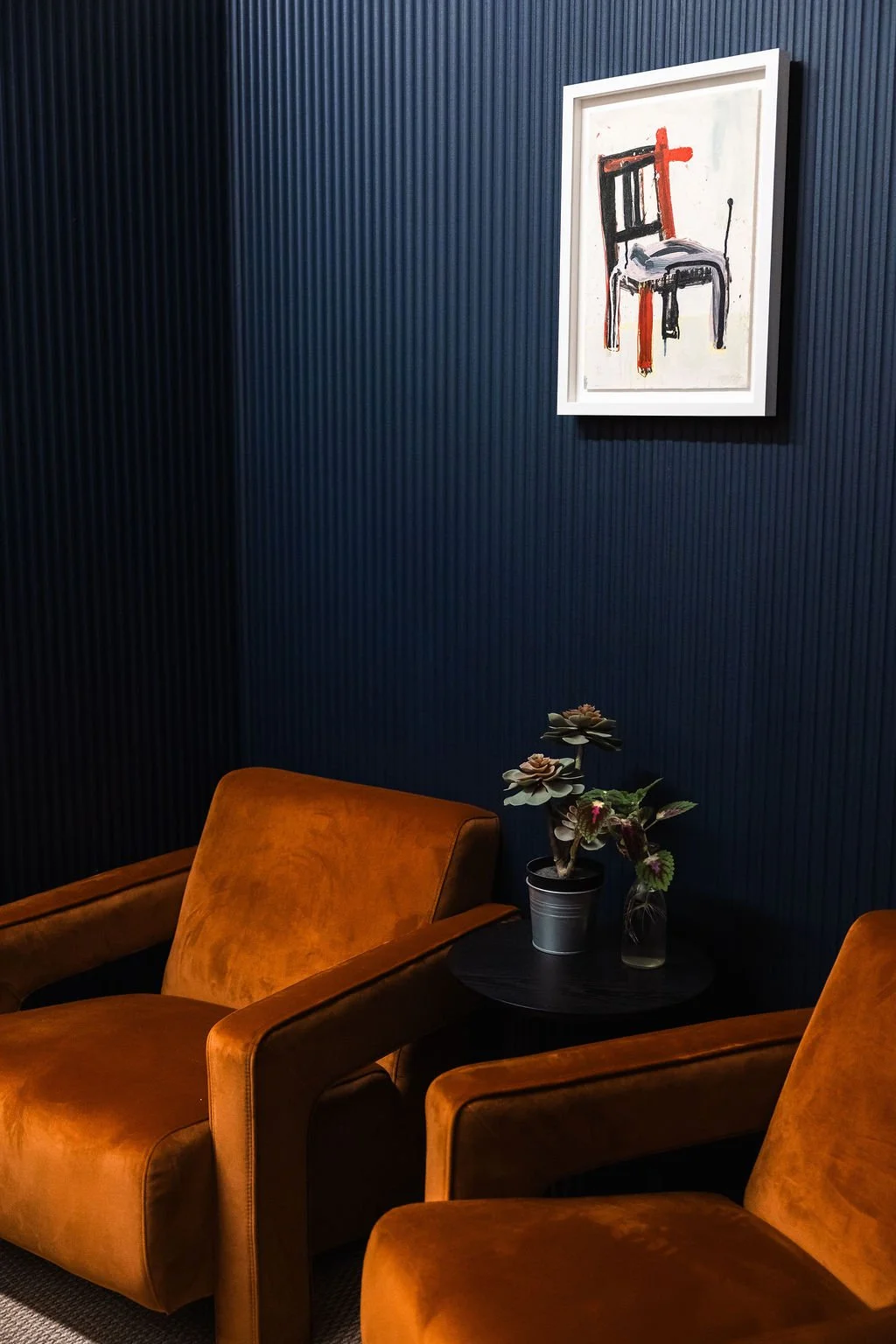 A cozy interior corner with two brown velvet armchairs, a small black side table with potted plants, a dark blue textured wall, and a white framed abstract painting with black, red, and gray brushstrokes.