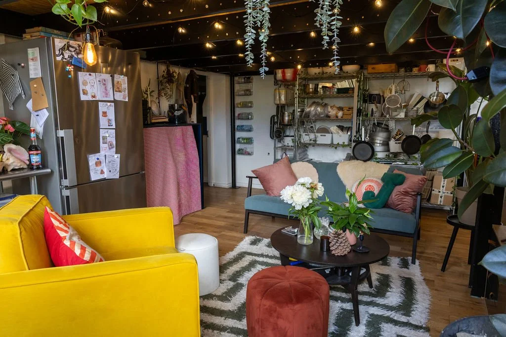 A cozy and colorful living room with a yellow couch, a blue sofa, and a wooden coffee table decorated with flowers, plants, and decorative items. The room features hanging string lights, a metal shelving unit with kitchenware, and a stainless steel refrigerator with children's drawings on it. The space is decorated with pillows, a patterned rug, and leafy plants.