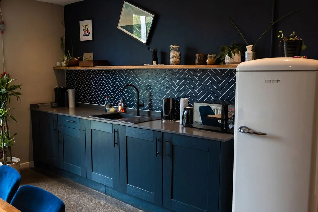 Modern kitchen with dark blue cabinets, a black backsplash, and a white vintage Gorenje refrigerator. Countertop has a sink, appliances, and decorative items. Wall has artwork and a mirror.