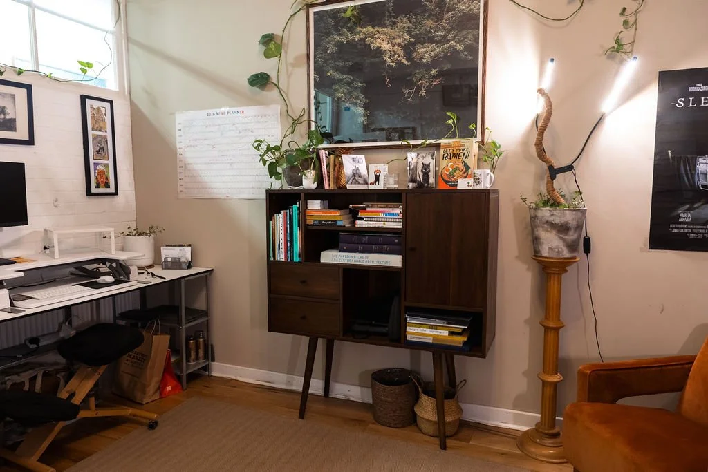 A cozy home office with a wooden desk, multiple computer monitors, and black office chairs. The wall features framed artwork, a wall calendar, and a large mirror reflecting greenery. Potted plants and decorative baskets are on a mid-century modern bookshelf and a tall plant stand. There's a brown chair in the foreground.