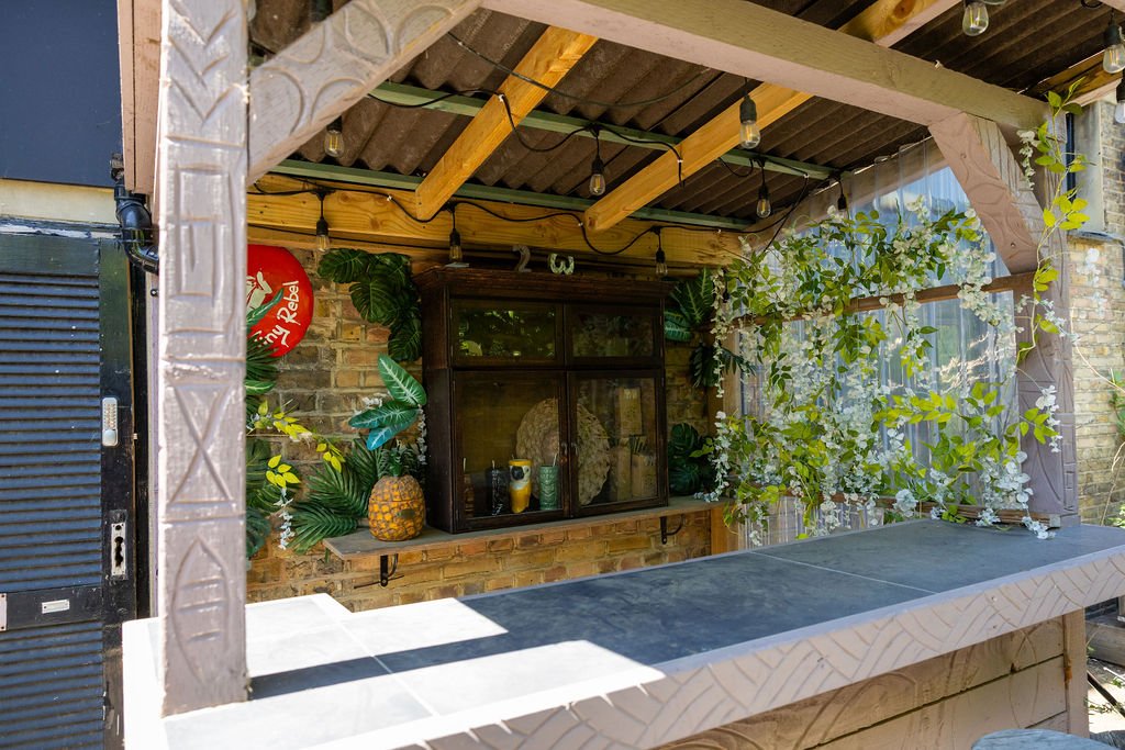 Outdoor bar with a brick wall background, decorated with green plants, a pineapple, and glassware, under a wooden and metal roof with string lights.
