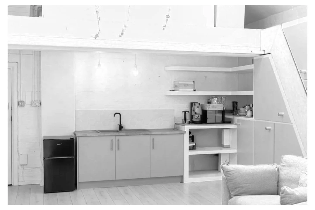 Small kitchen area with a mini fridge, a sink, and open shelves holding small appliances and kitchen items. Part of a living area with a sofa is visible in the foreground.