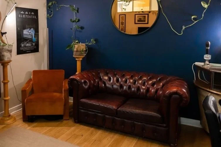 Living room with a brown armchair, a dark brown leather couch, a round mirror on a blue wall, and potted plants on wooden stands.