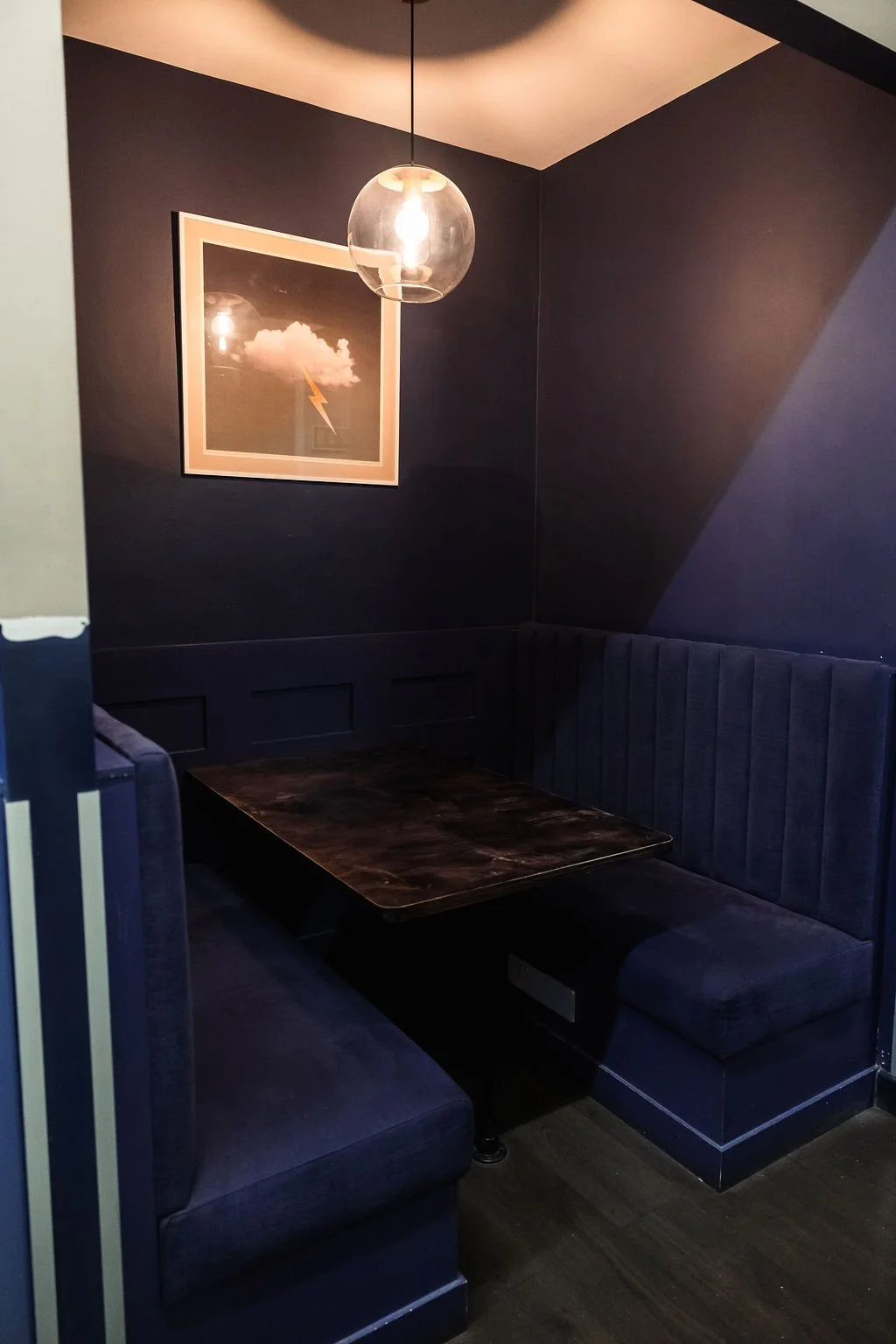 A cozy corner booth with dark blue upholstered seating, a wooden table, a framed picture of a cloud with a lightning bolt, and a hanging glass light fixture on a dark wall.
