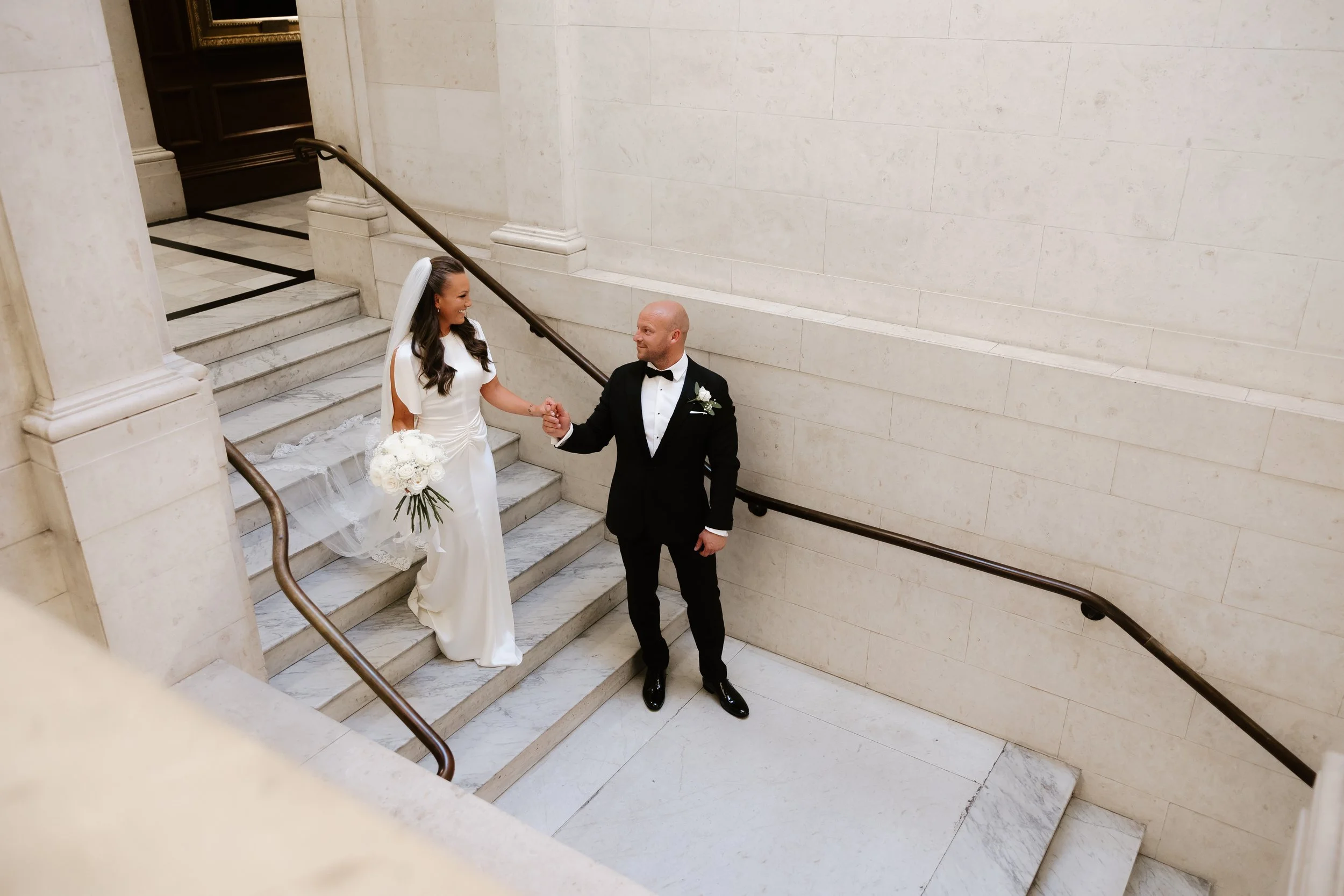 Bride in white dress with long veil and groom in tuxedo, walking hand in hand down the steps inside the Old Marylebone Town Hall in London. The groom is leading the bride down the white marble  steps. 