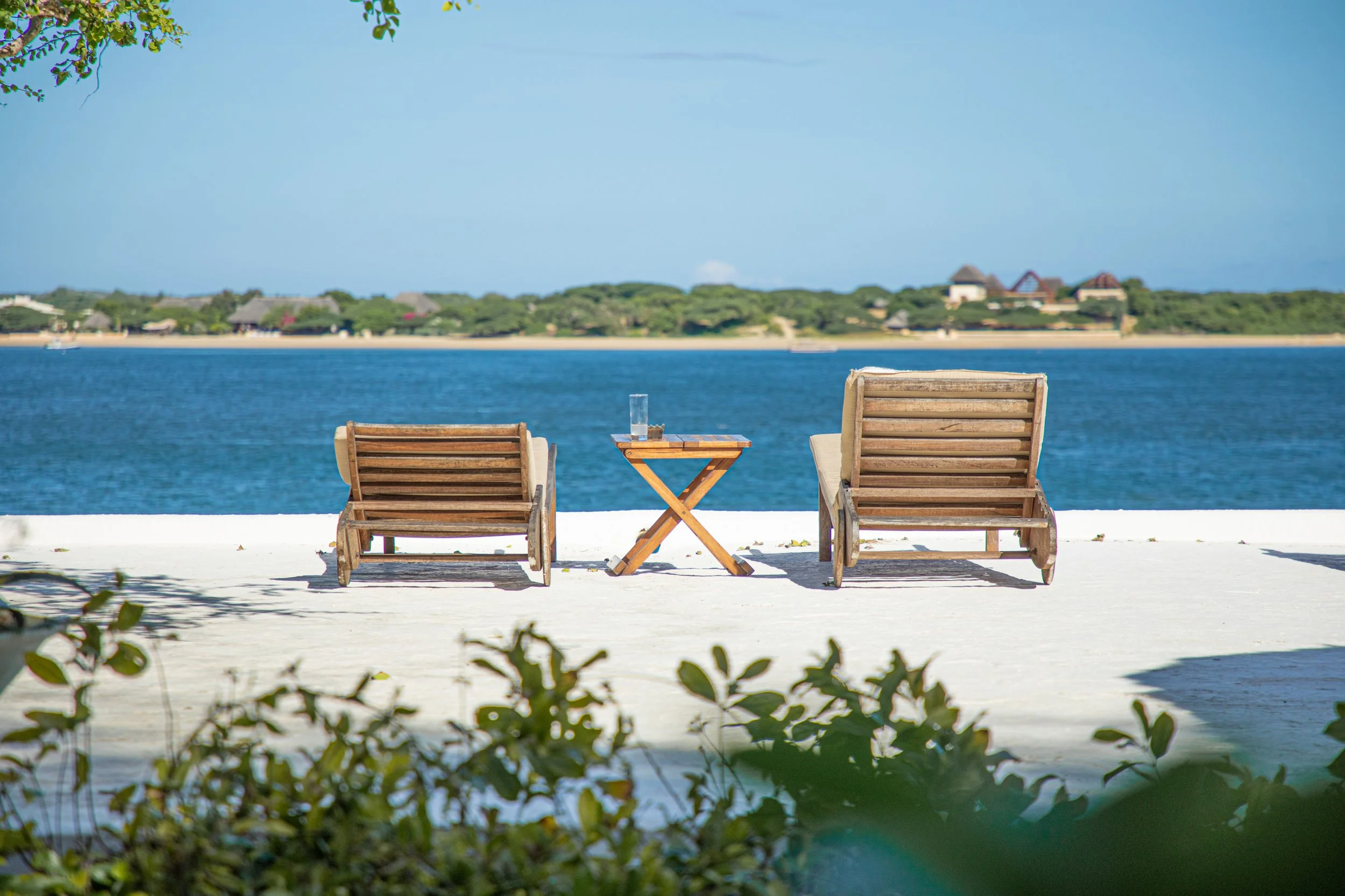 Two wooden lounge chairs with beige cushions facing a body of water with houses and greenery in the background, a small wooden table with a glass of water and a small item between them on the white sand beach.