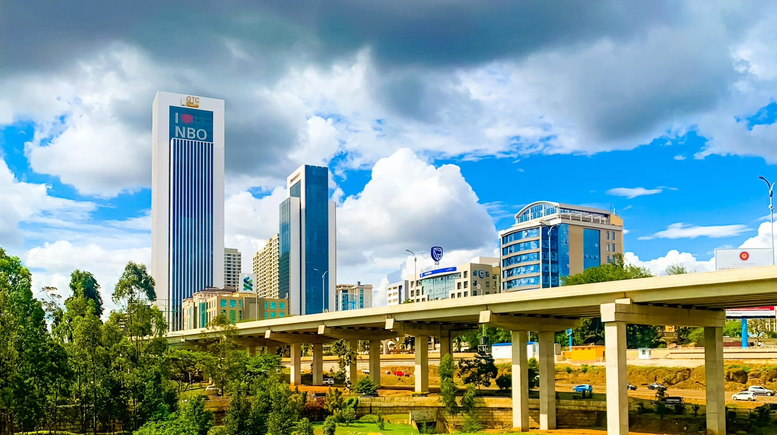 City skyline with modern high-rise buildings under a partly cloudy sky, with a highway in the foreground and green trees below.