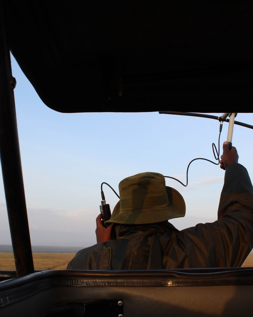 A person wearing a hat and jacket sitting in a vehicle field, holding radio equipment, with open sky and grassy landscape in the background.
