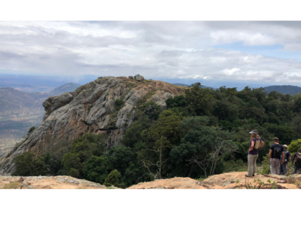 Hikers walking on a rocky trail overlooking a lush green valley with large rocks on the mountainside and cloudy sky above.