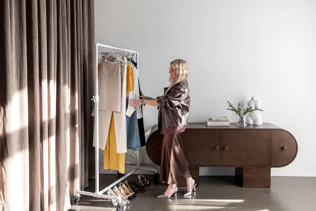 Kathryn Snow, personal stylist, fashion expert, and wardrobe consultant in Grand Rapids, Michigan, shopping for clothes in a minimalist room with a clothing rack, a dark wooden sideboard, and a white wall.