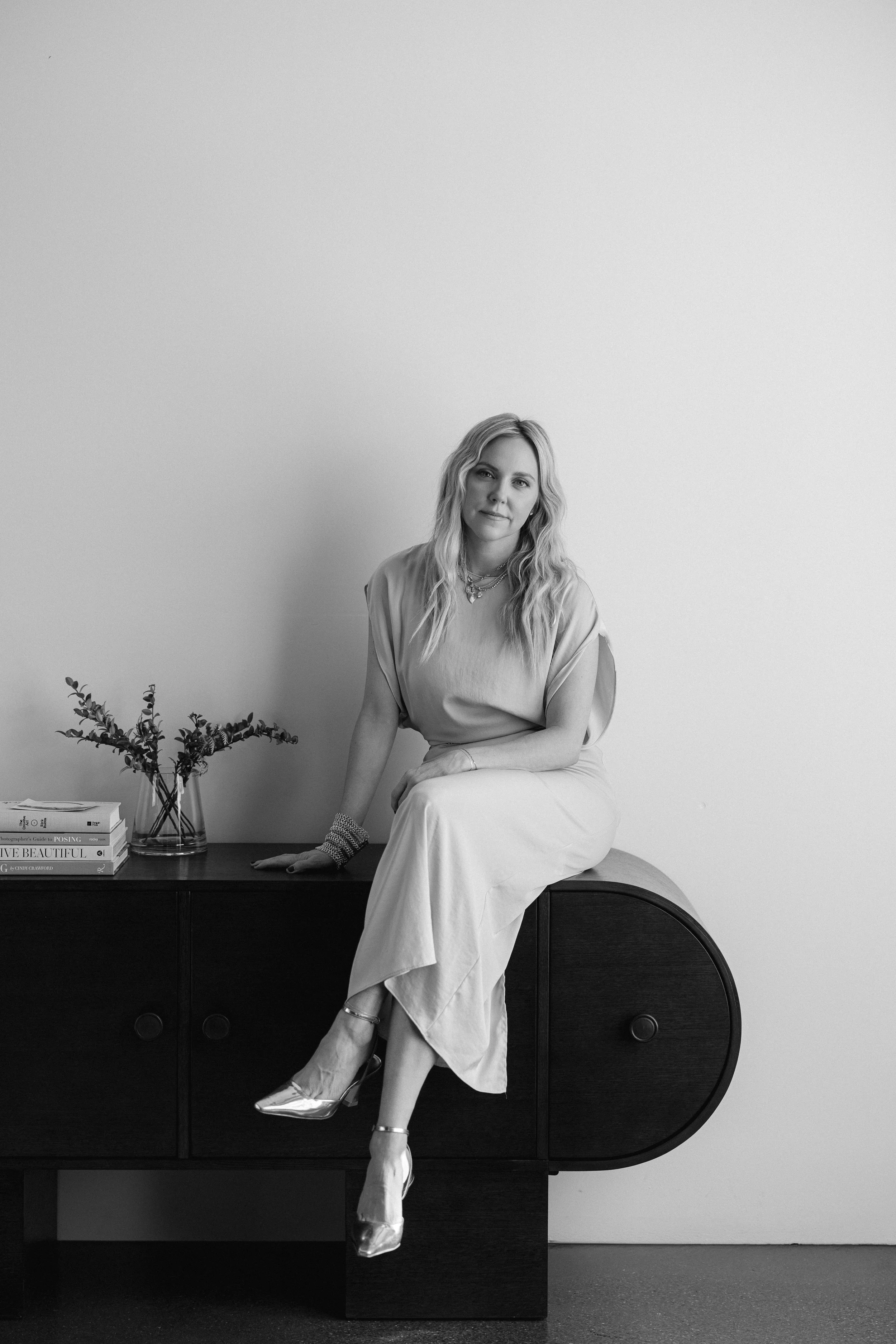 Kathryn Snow, personal stylist, fashion expert, and wardrobe consultant in Grand Rapids, Michigan, sitting on a wooden cabinet, with a plant and magazines on top, against a plain wall.