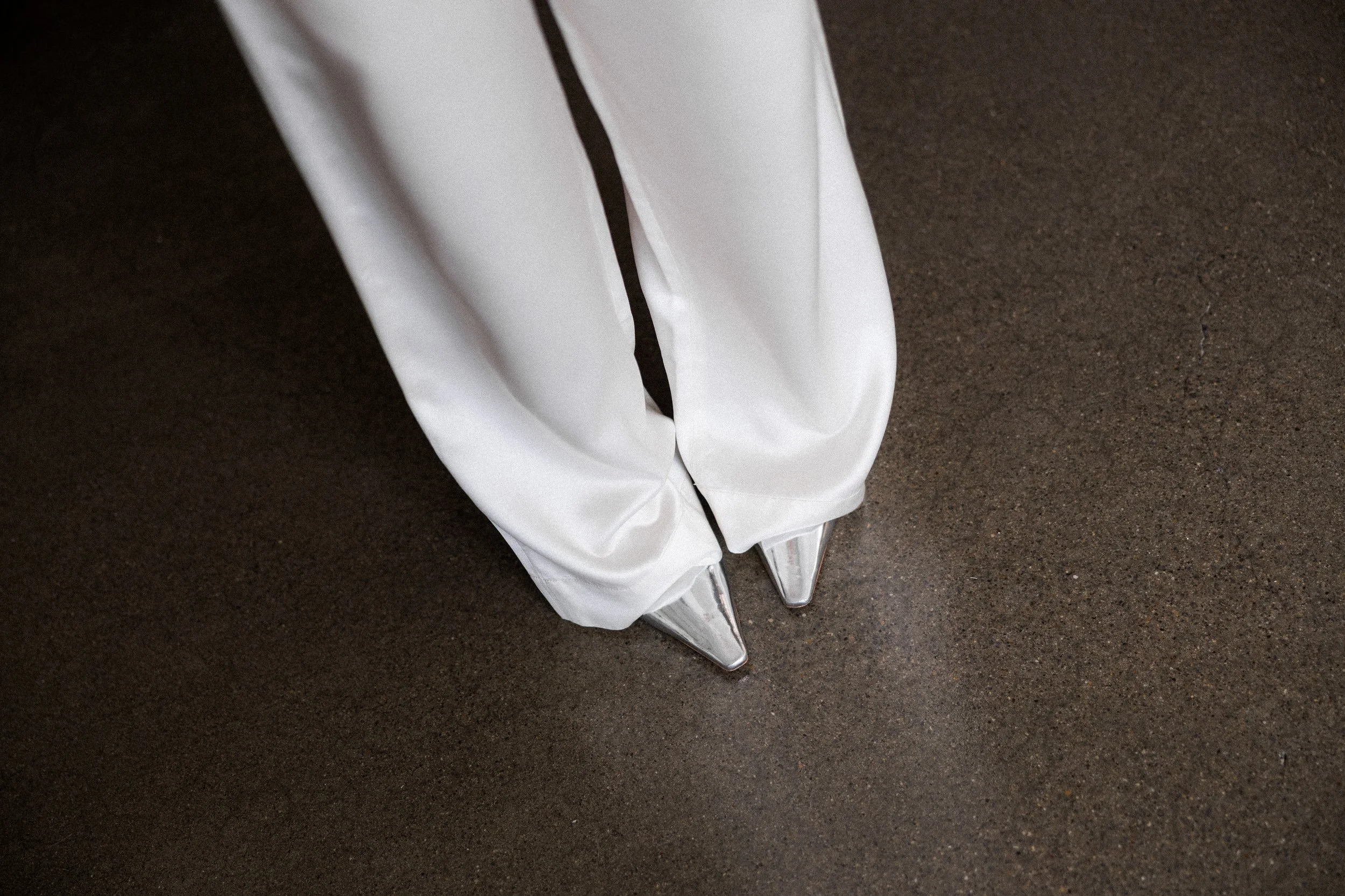 Close-up of the bottom of white pants with silver pointed toe shoes on a dark textured floor. These shoes belong to Kathryn Snow, personal stylist, fashion expert, and wardrobe consultant in Grand Rapids, Michigan