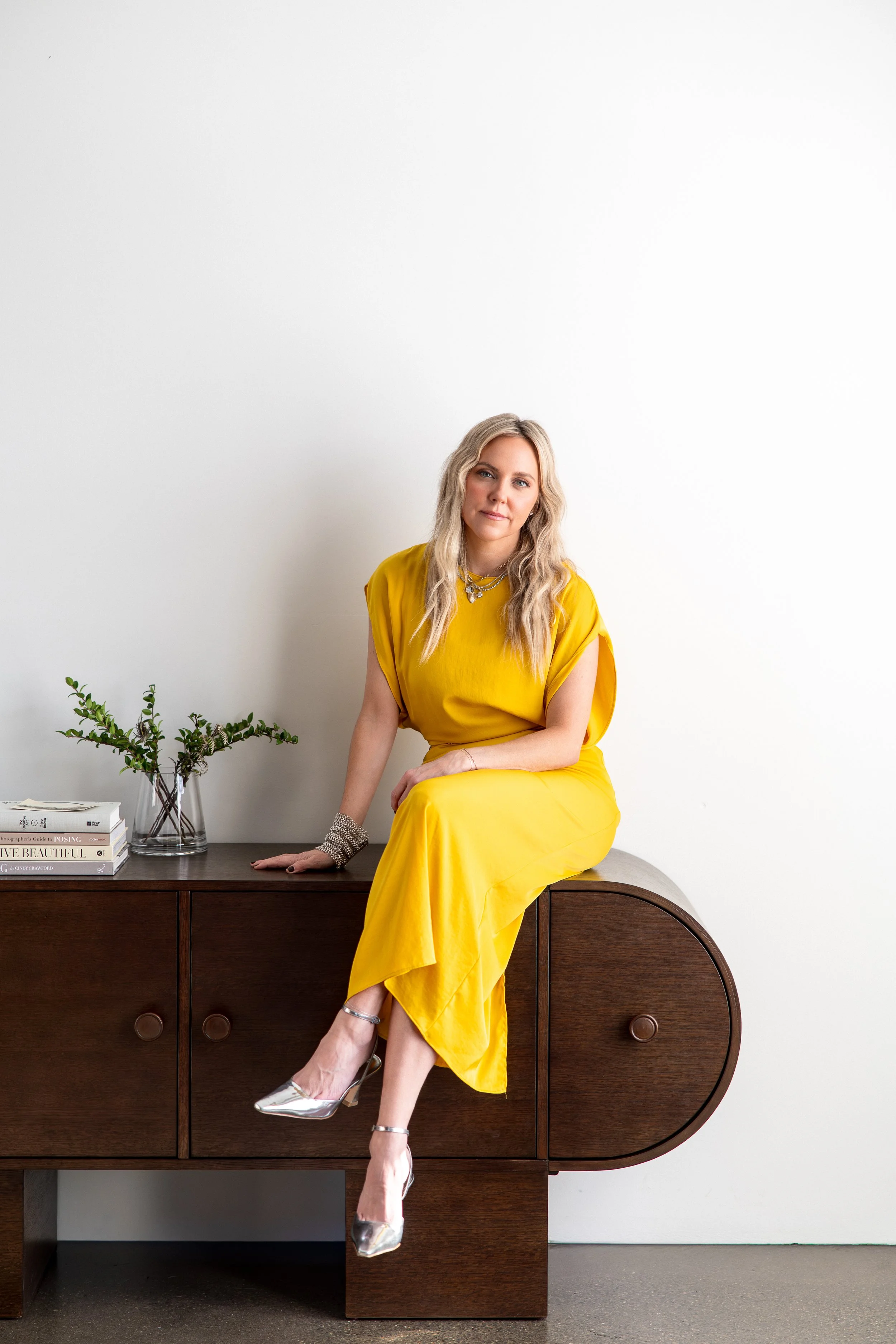 Kathryn Snow, personal stylist, fashion expert, and wardrobe consultant in Grand Rapids, Michigan sitting on a dark wooden sideboard with a white wall in the background, a small book, and a glass vase with green foliage.