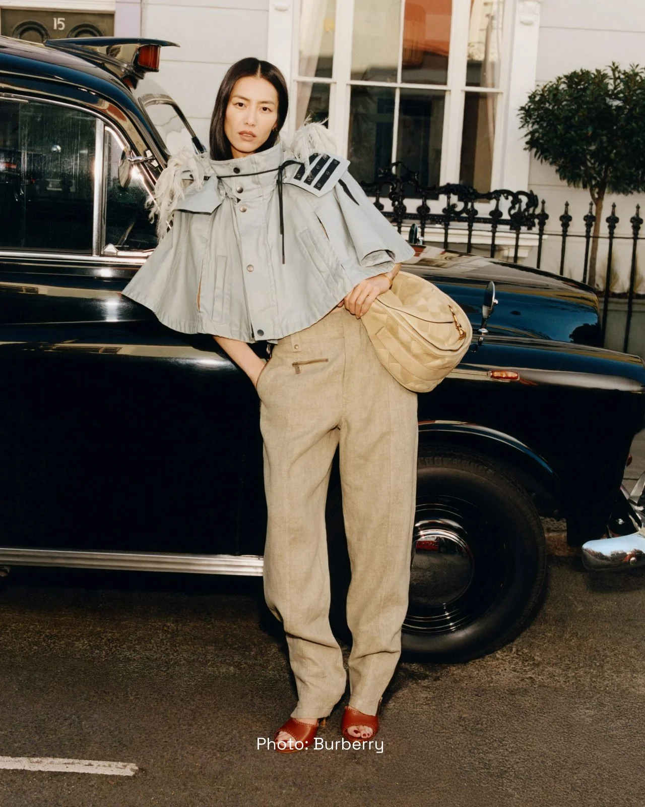 A woman standing next to a vintage black car, dressed in beige pants, a light gray cropped jacket with shoulder embellishments, carrying a beige quilted bag, wearing brown shoes, and posing outdoors in front of a white house with black iron fence and greenery.