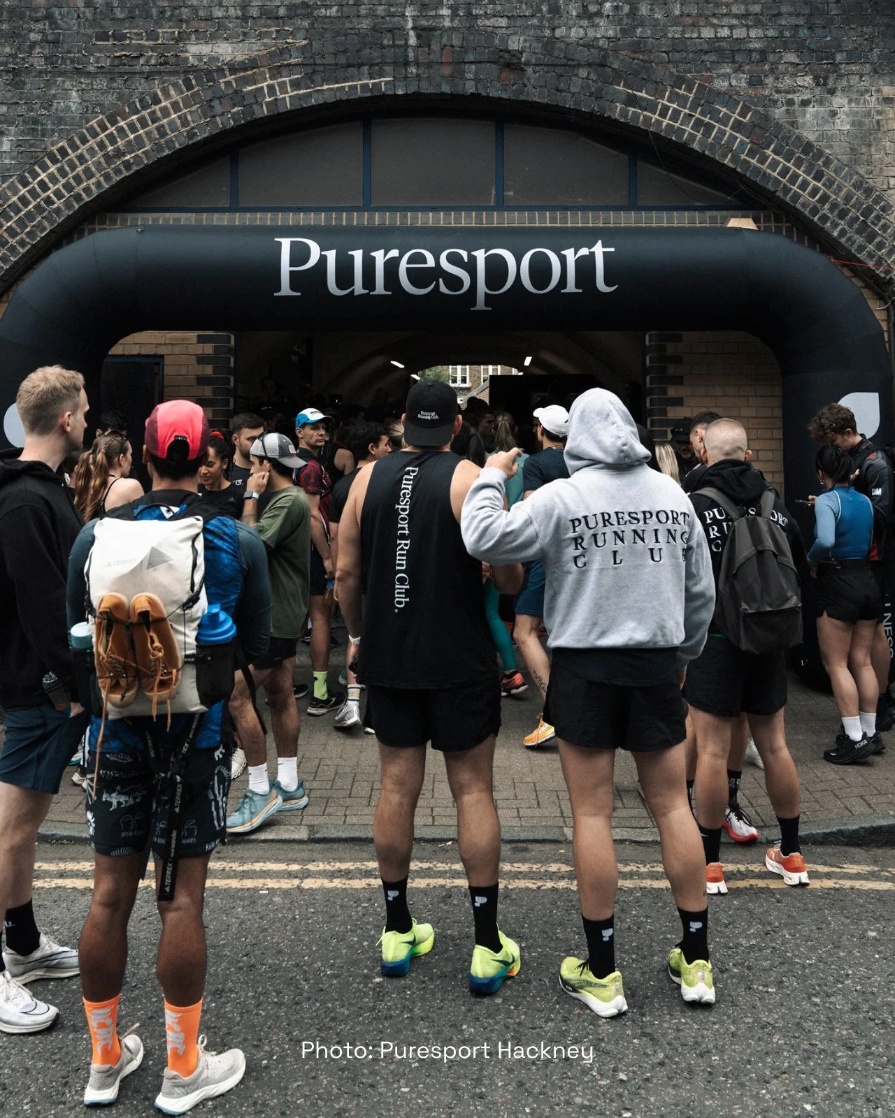 Group of runners gathered under Puresport archway before a race, wearing athletic gear and backpacks, in Hackney.
