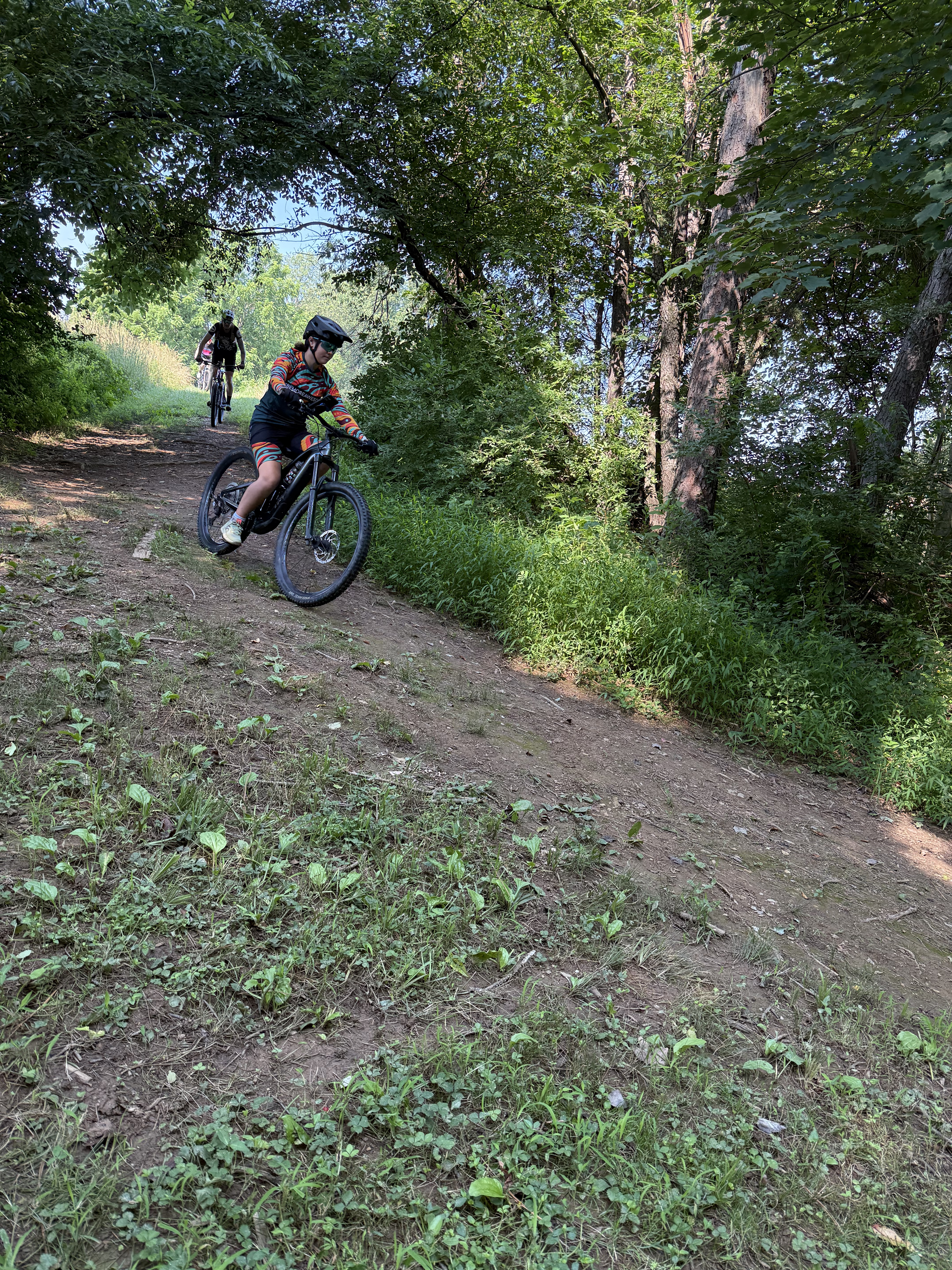 Two children riding mountain bikes on a dirt trail through a green, wooded area with trees and bushes.
