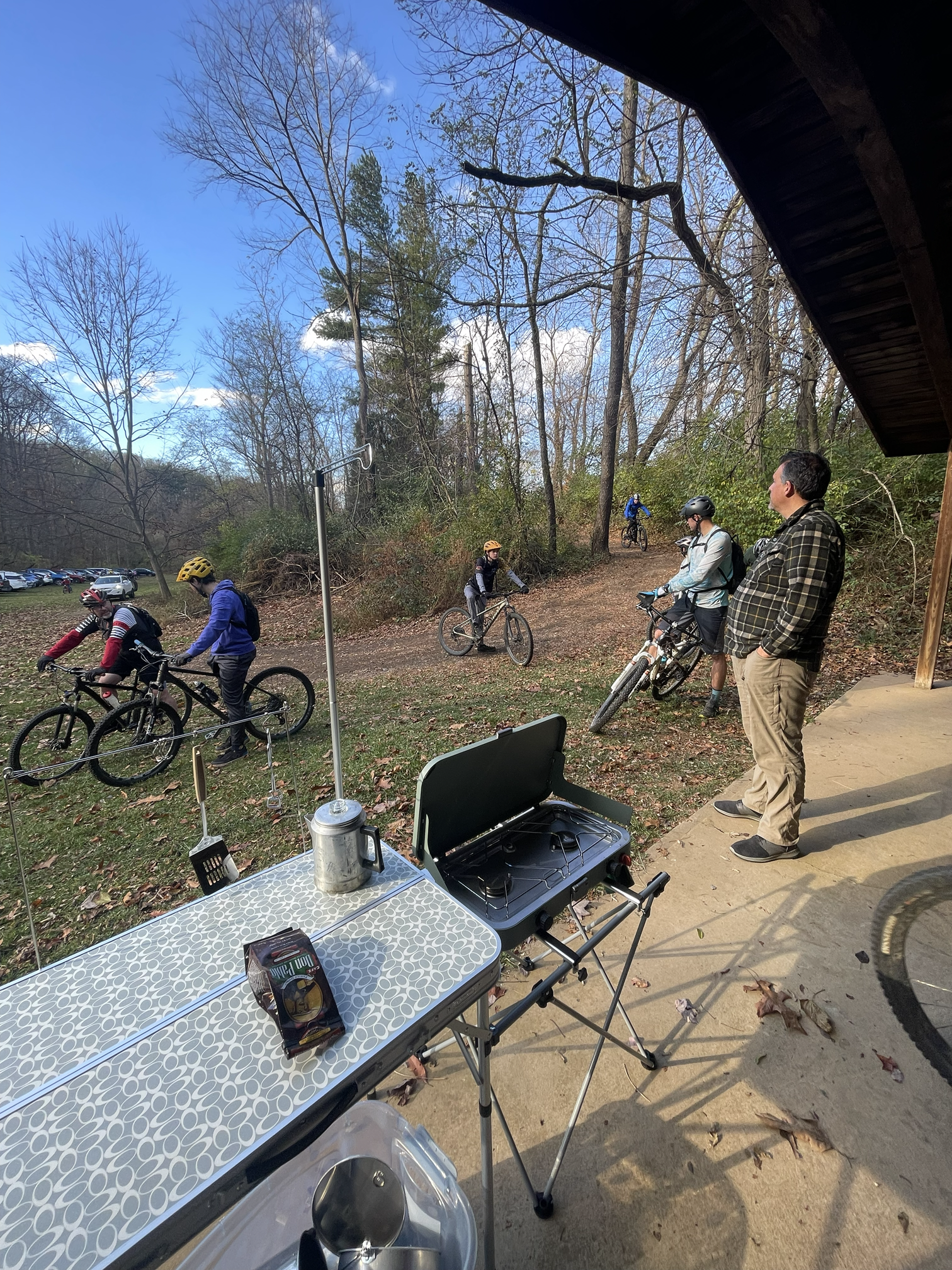 A group of people with bicycles gathered outside in a wooded area, some wearing helmets, with a table and grill set up in the foreground.