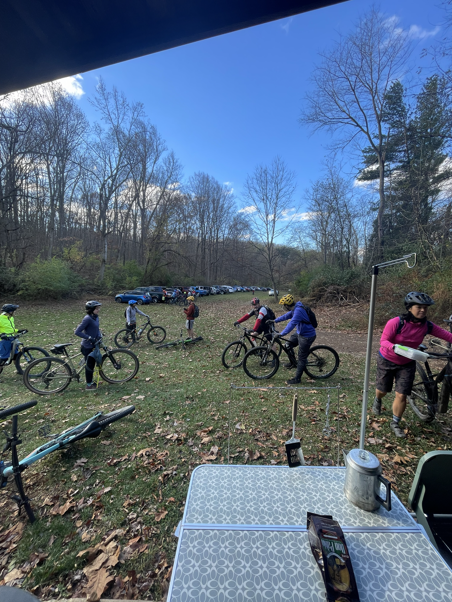 People in cycling gear gathered outdoors among trees, with parked cars in the background, under a partly cloudy sky.