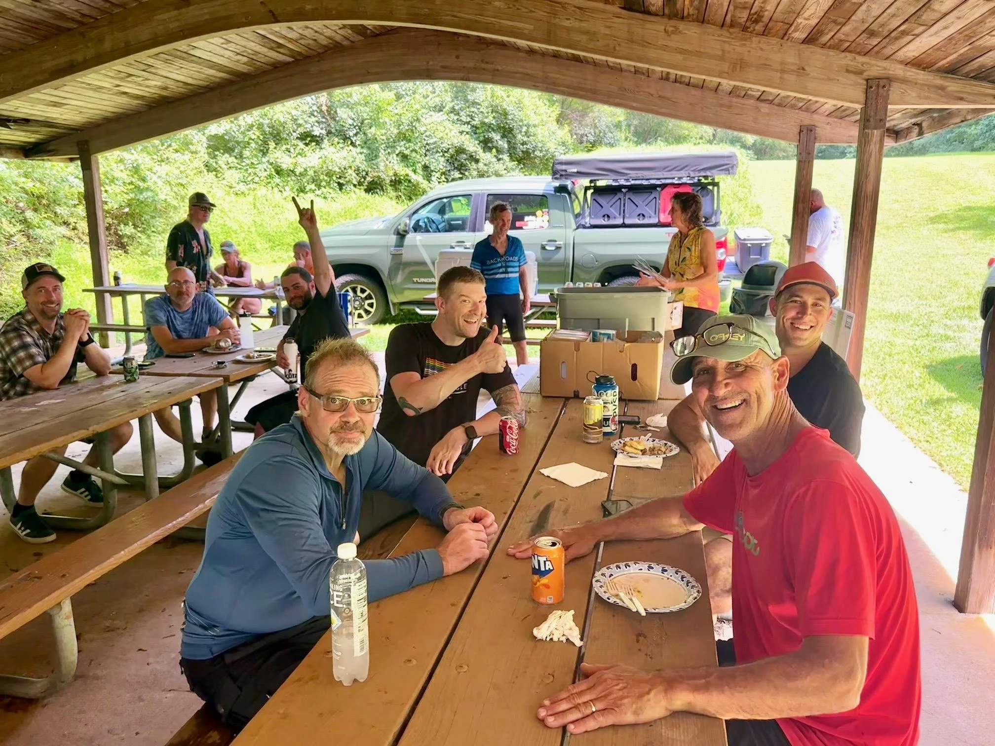 Group of smiling people gathered around picnic tables at an outdoor event, with some eating and others chatting under a wooden pavilion.