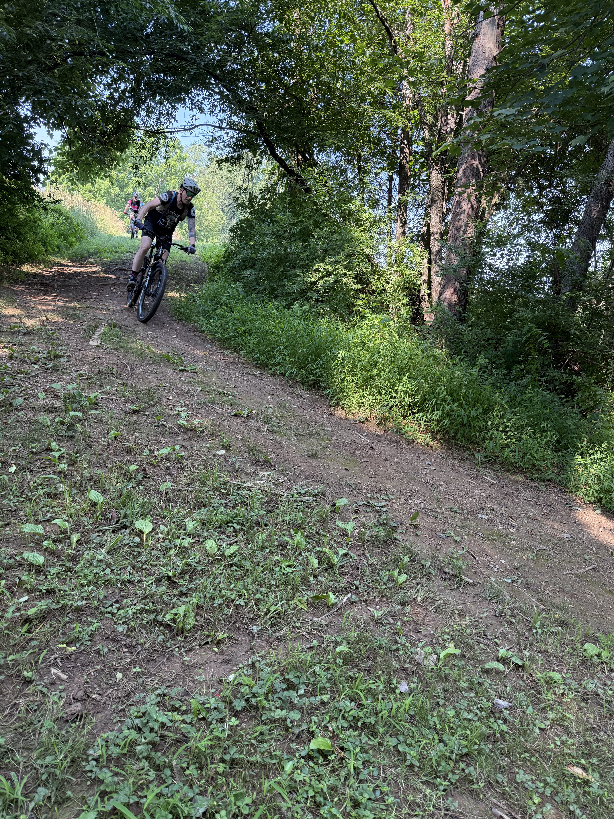 People mountain biking on a dirt trail through green trees in a wooded area on a sunny day.
