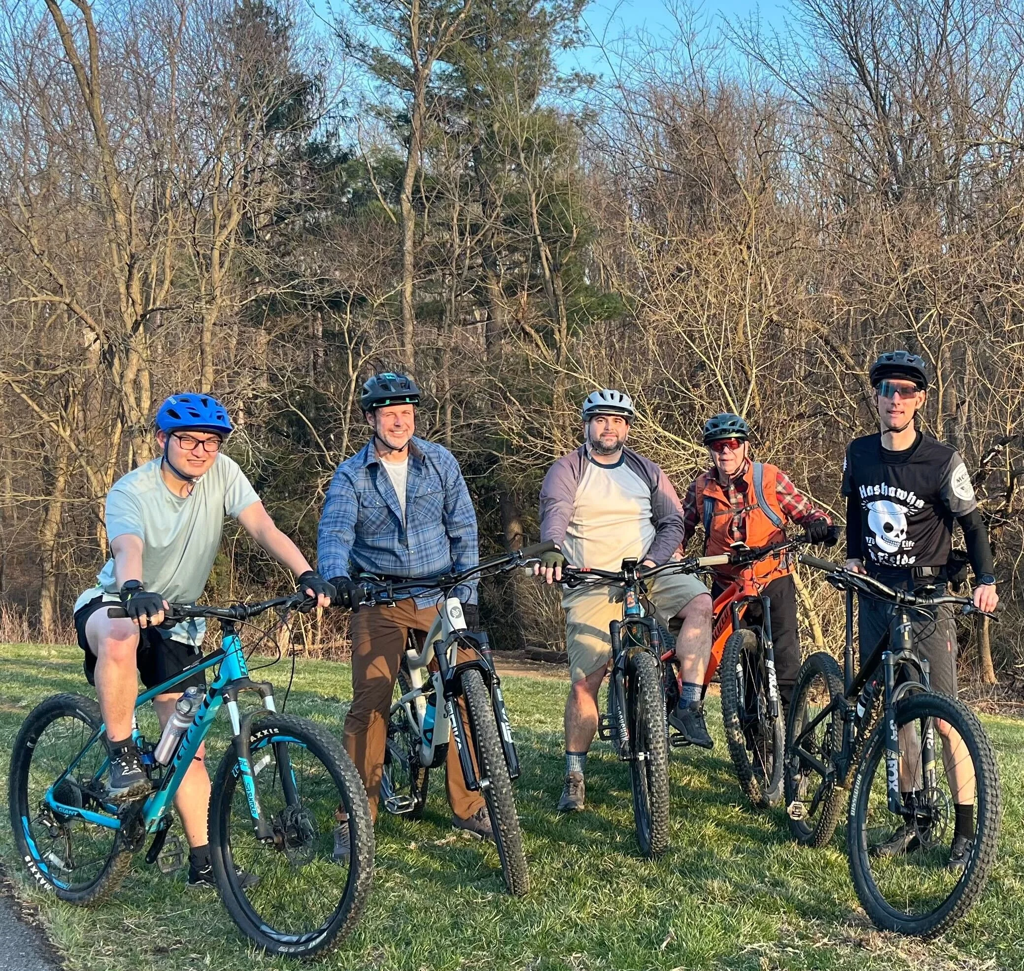 Group of five men with mountain bikes standing outdoors on grassy area, wearing helmets and casual clothing, with leafless trees in the background.