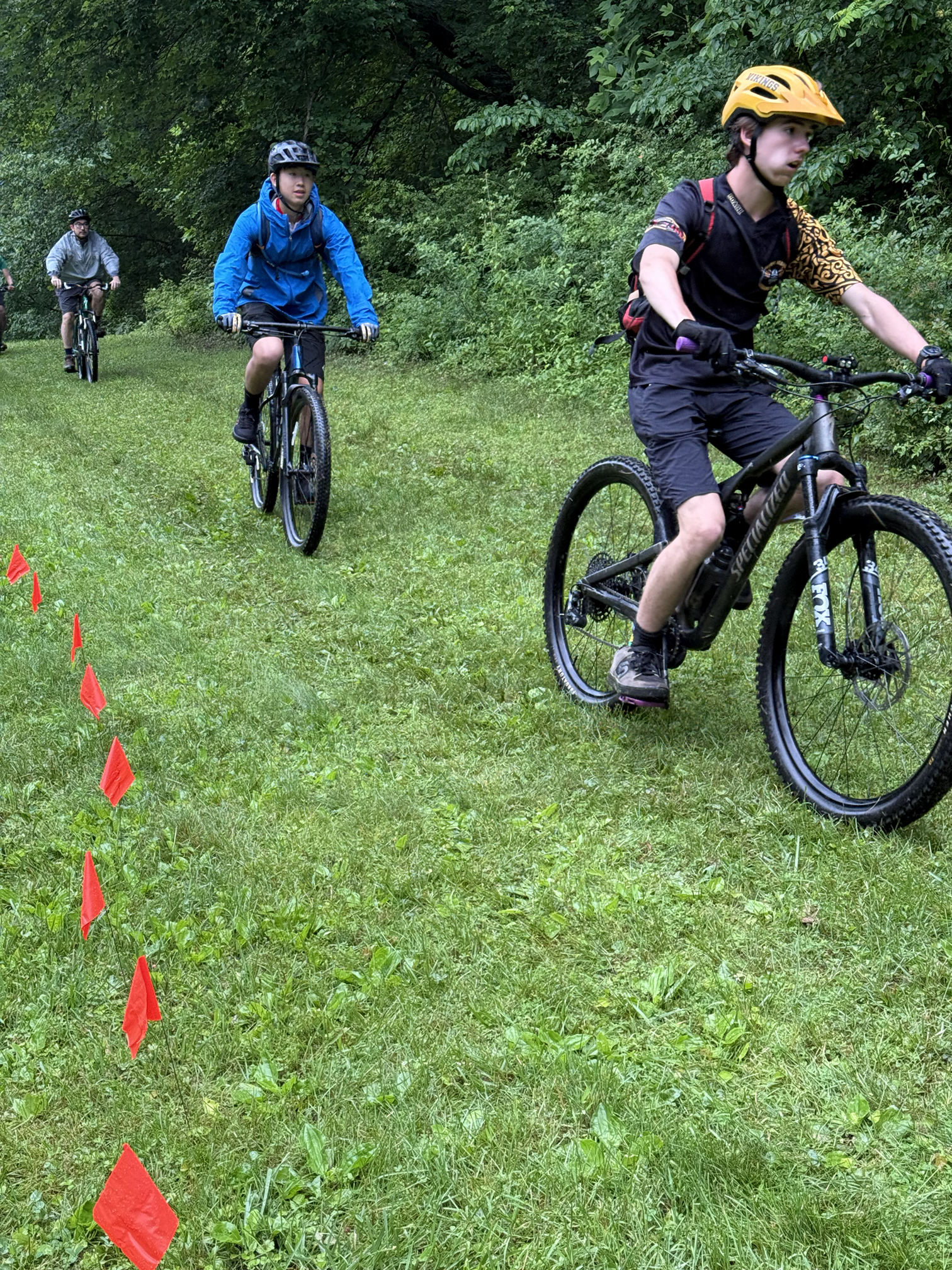 Three young men riding mountain bikes along a grassy trail surrounded by green trees. They are wearing helmets, and the trail is marked with small red flags on the left side.