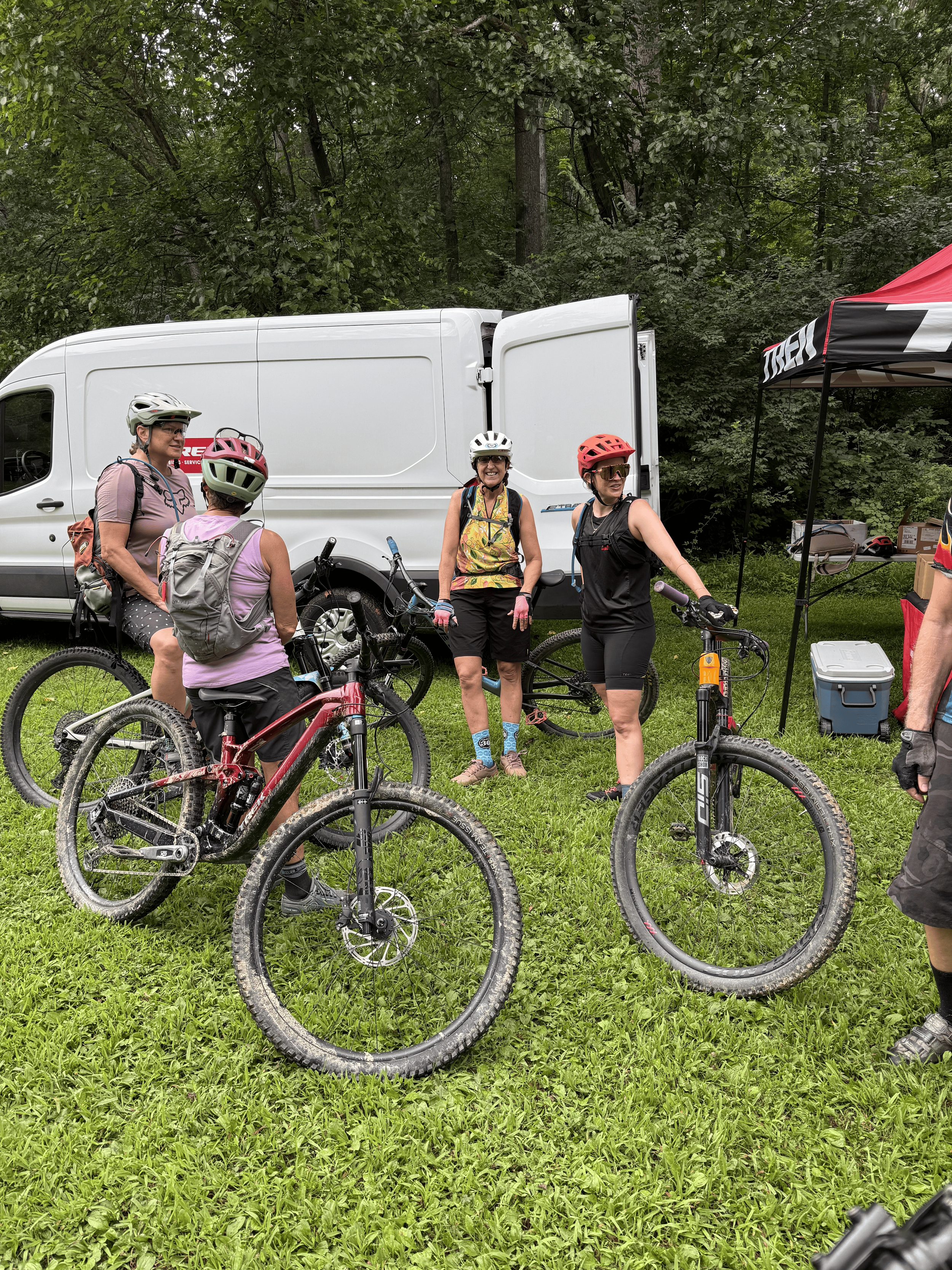 Group of women in cycling helmets and gear standing with mountain bikes outdoors near a white van and a red and black tent.