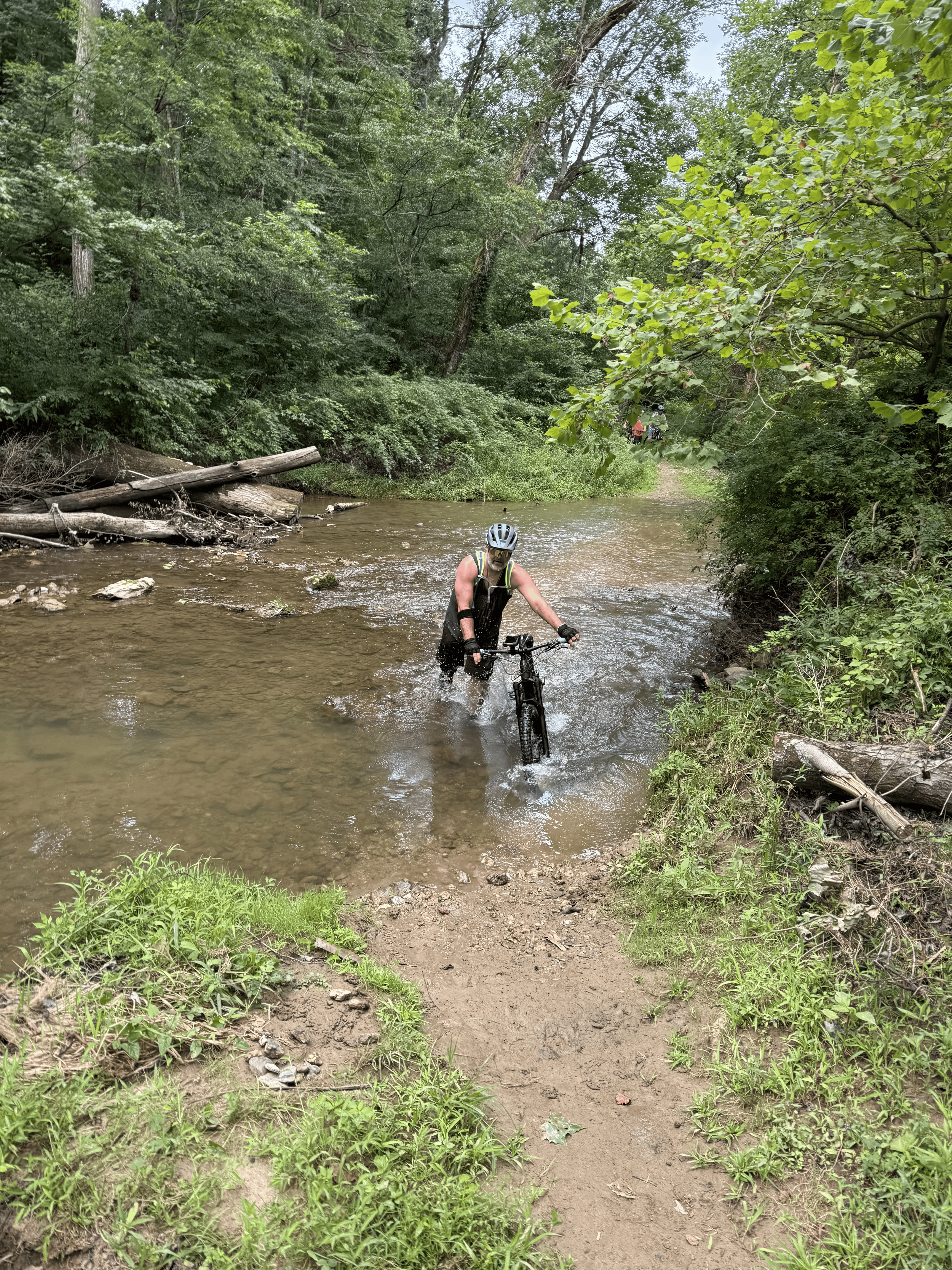 A person riding a mountain bike through a shallow stream in a lush green forest, wearing a helmet and sleeveless shirt.