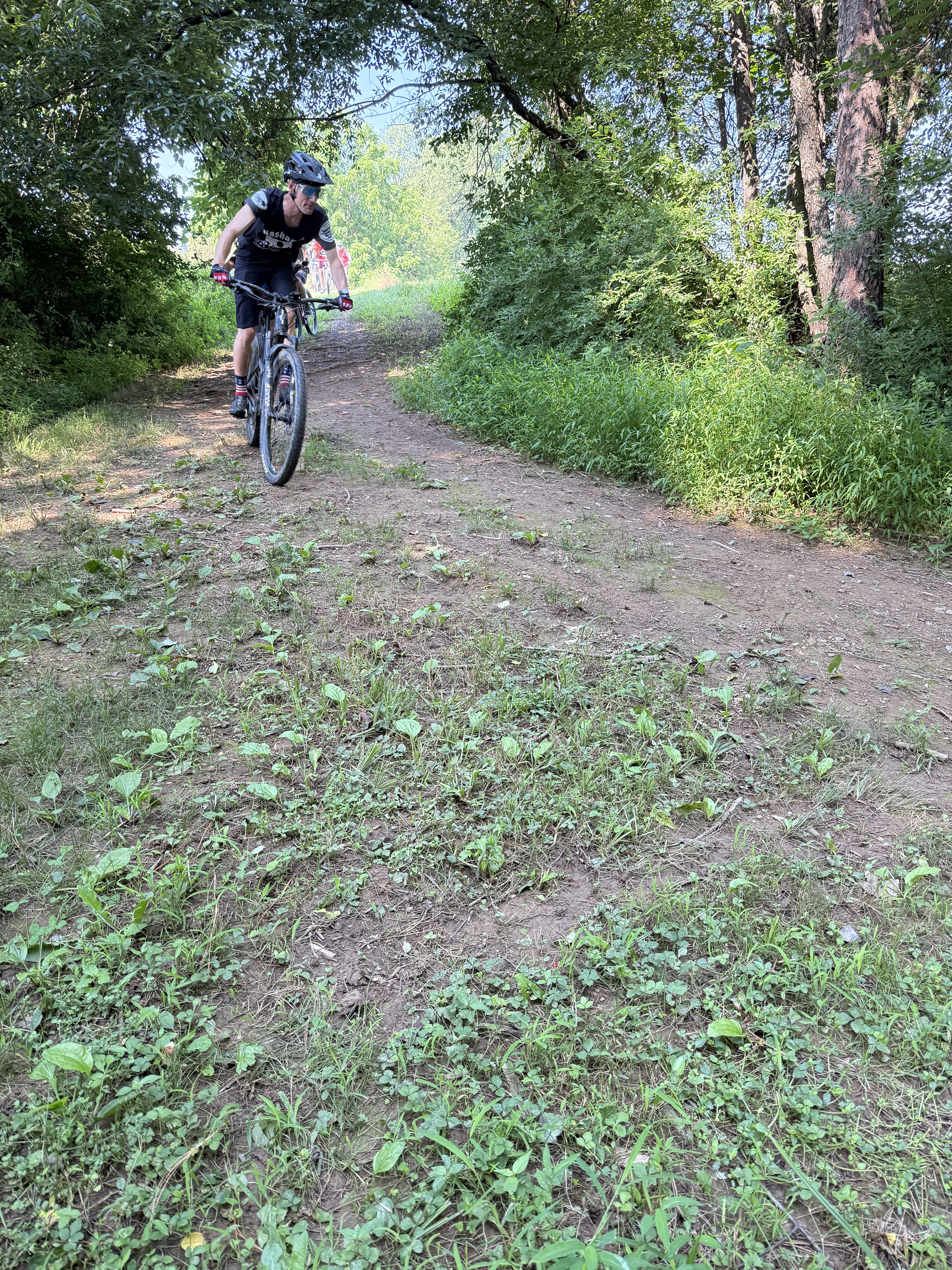 A man wearing a black helmet, sunglasses, and black cycling attire is riding a mountain bike on a dirt trail through a lush green forest.