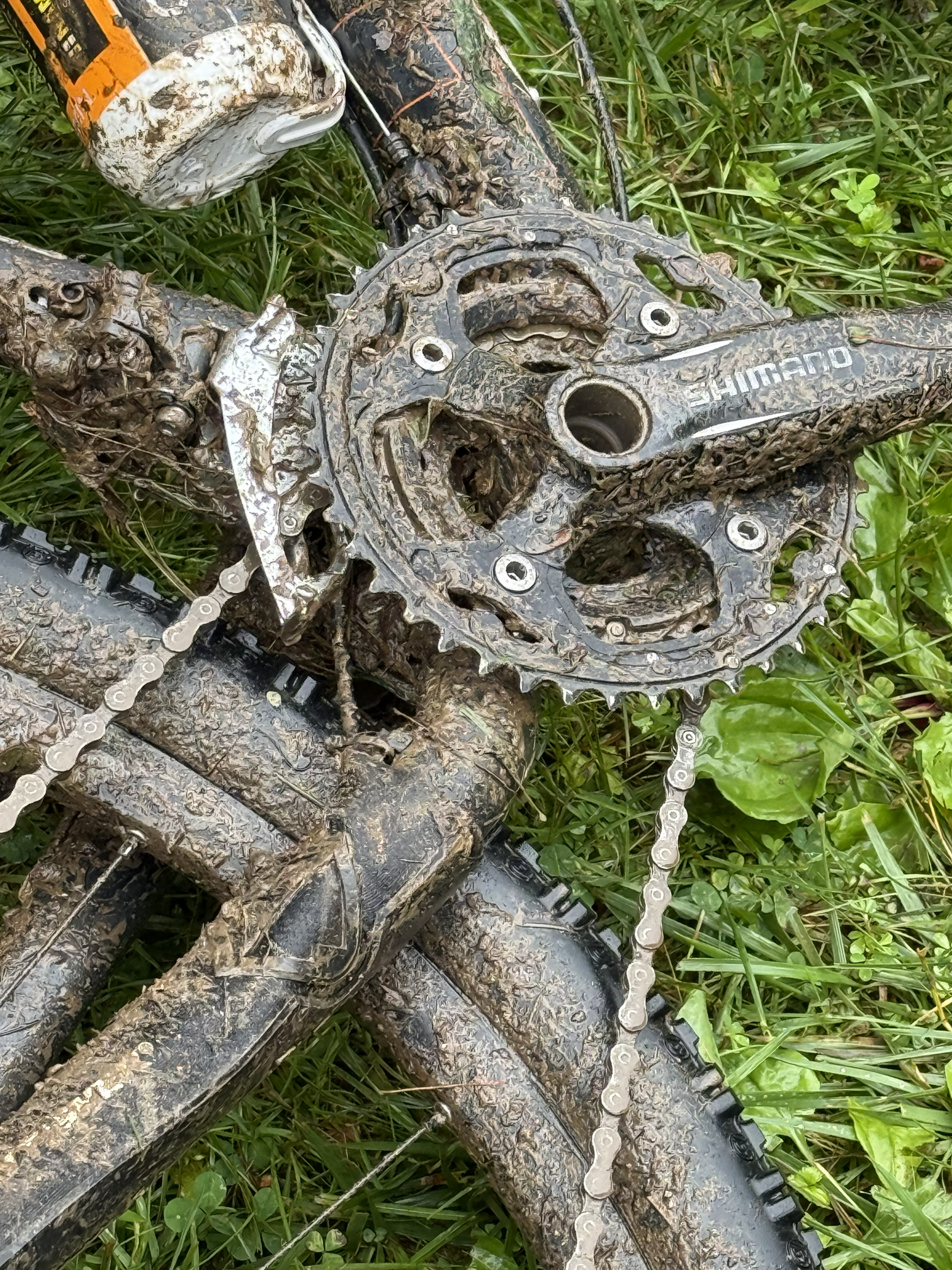 A muddy mountain bike lying on the grass, showing the chain, gears, and crankset covered in mud.
