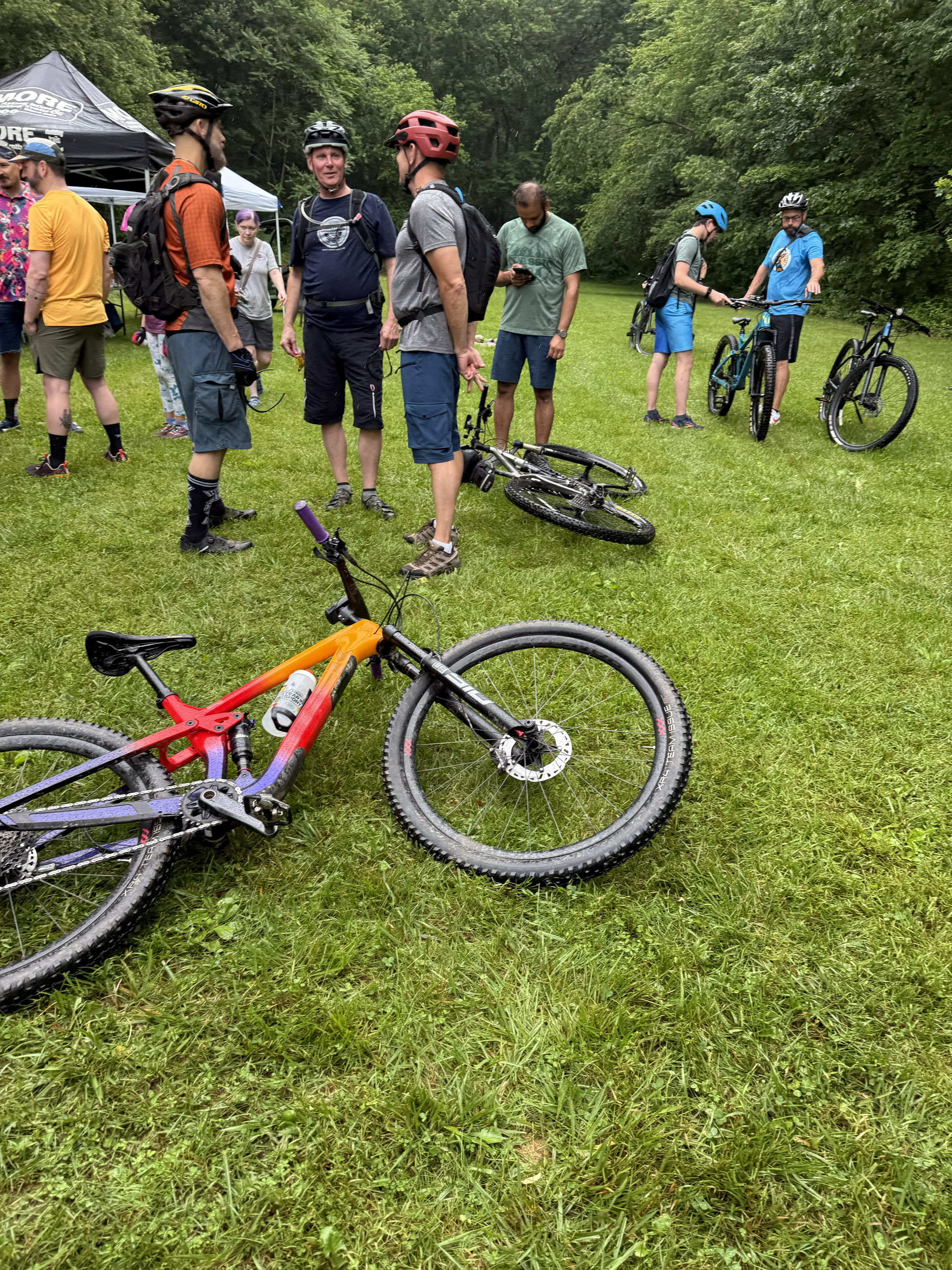 A group of people with bicycles gathered on a grassy field in a wooded area, some wearing helmets and casual outdoor clothing, engaging in conversation.