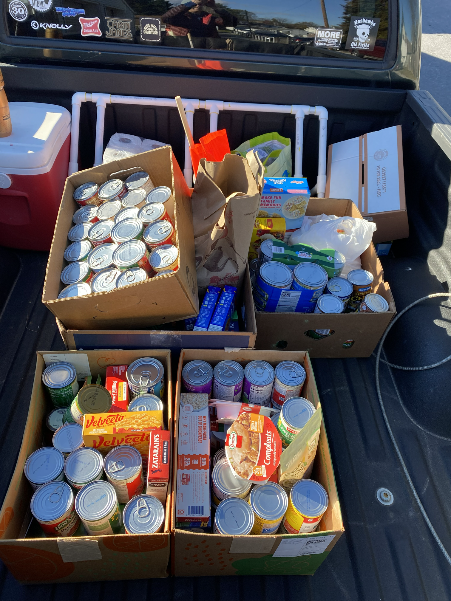 The image shows the bed of a pickup truck loaded with boxes of canned foods, instant noodles, and other non-perishable items for donation or distribution.