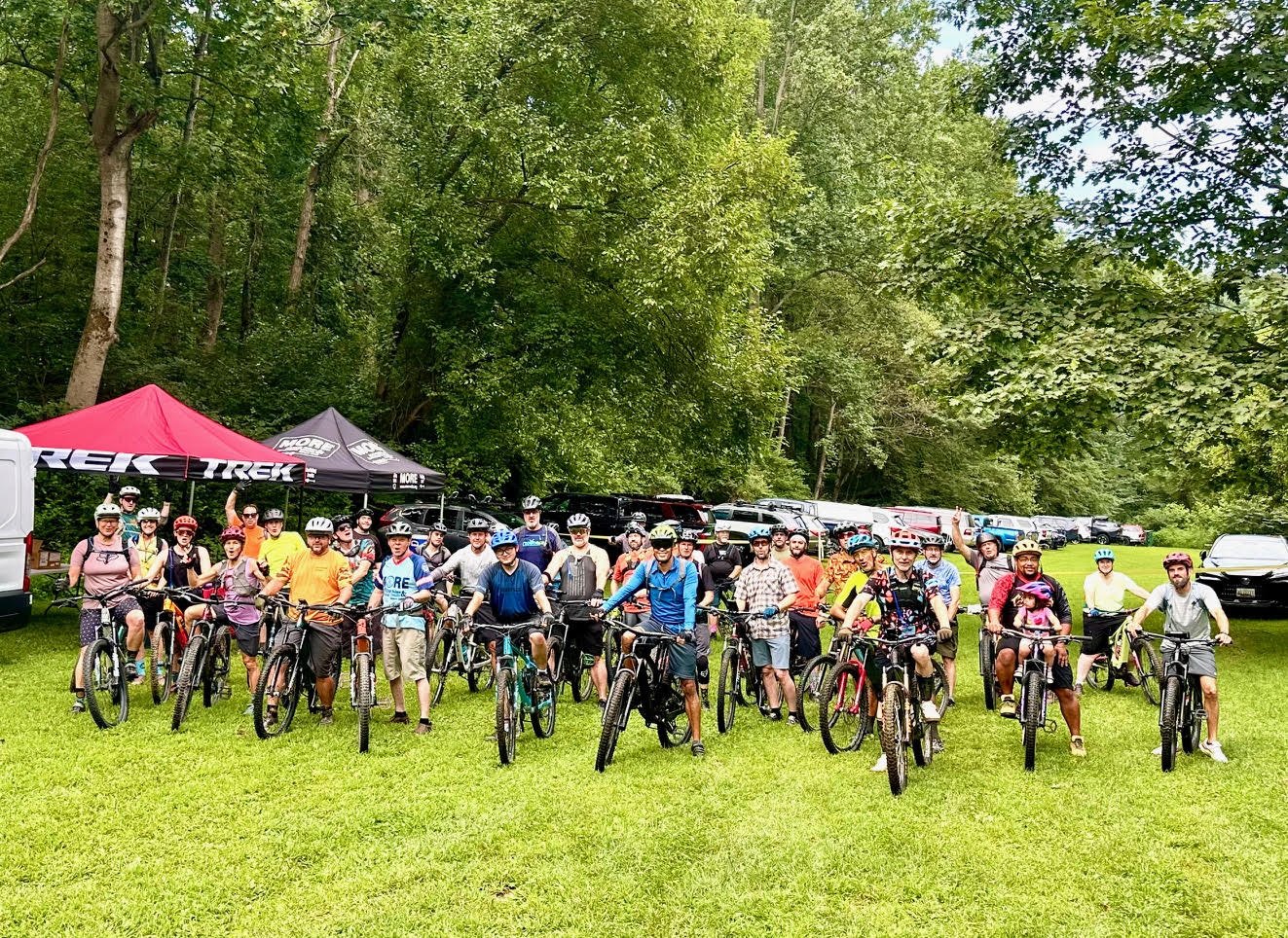 Group of people with bicycles outdoors in a grassy area with trees, tents, and parked cars in the background.