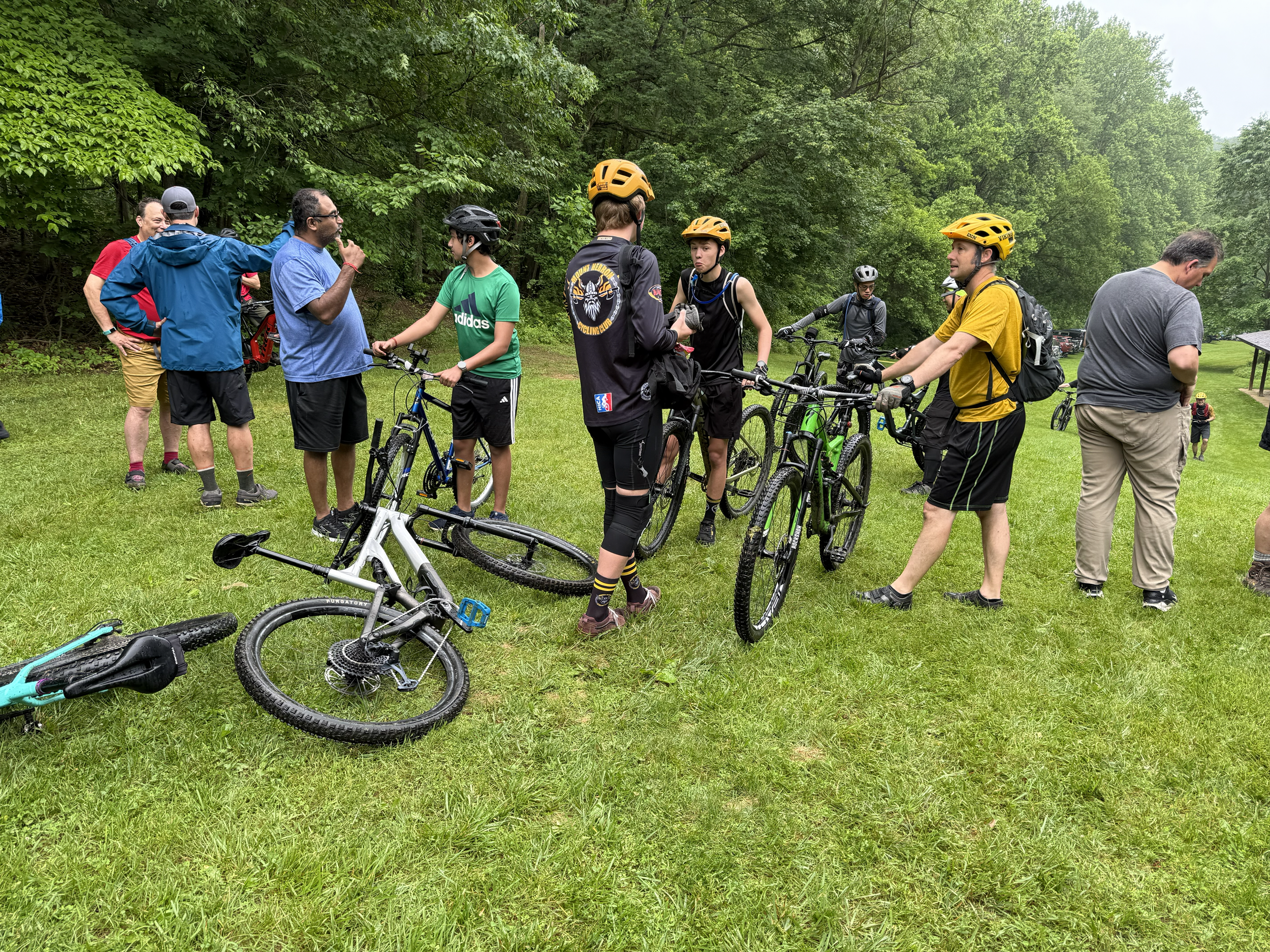 Several people, some wearing helmets, are gathered in a grassy area near a wooded area. They have bicycles with them, and some are talking while others are preparing to ride or taking a break.