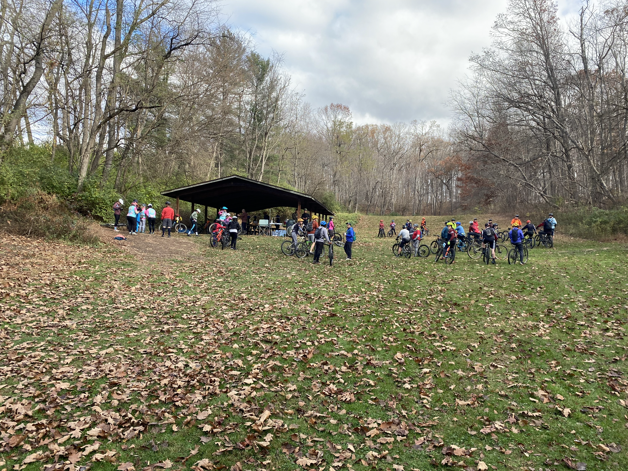 A group of people, many with bicycles, gathered under a shelter in a park surrounded by leafless trees, during a cloudy day.