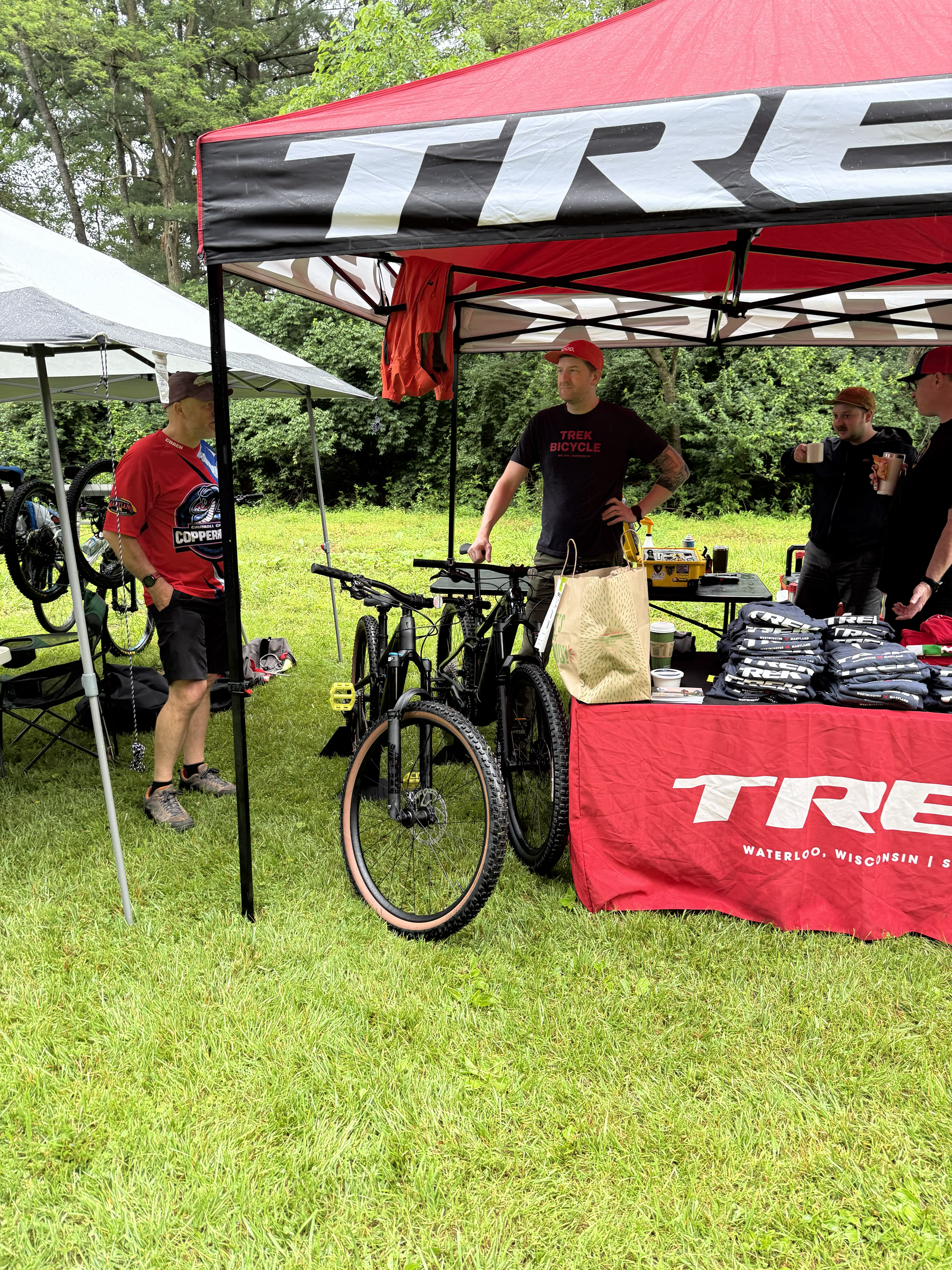 Group of men at an outdoor event under a red and white canopy, with cycling gear and merchandise on display, surrounded by green trees and grass.
