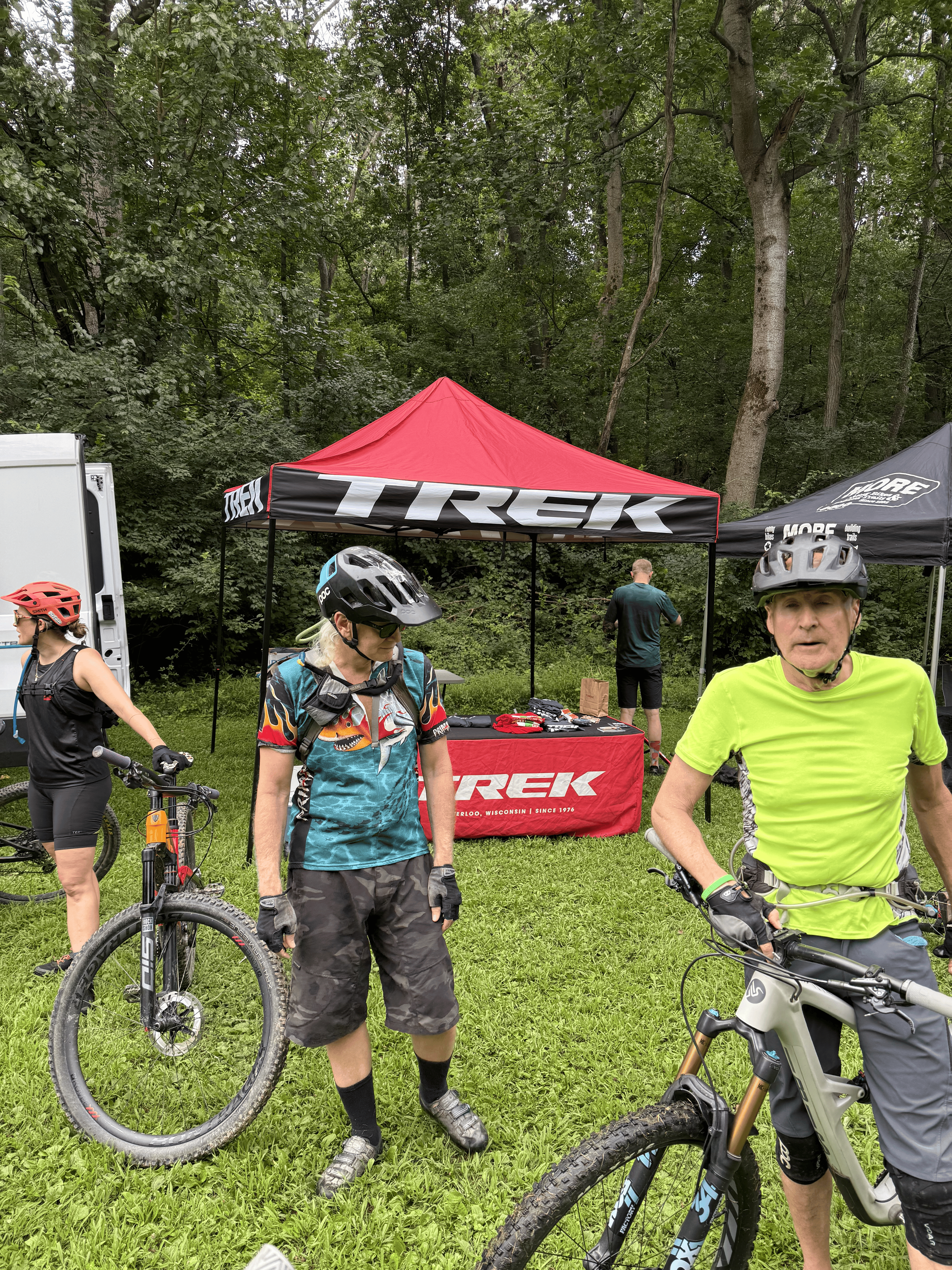 Group of cyclists in a wooded area near a red canopy tent with Trek branding, wearing helmets and riding mountain bikes.