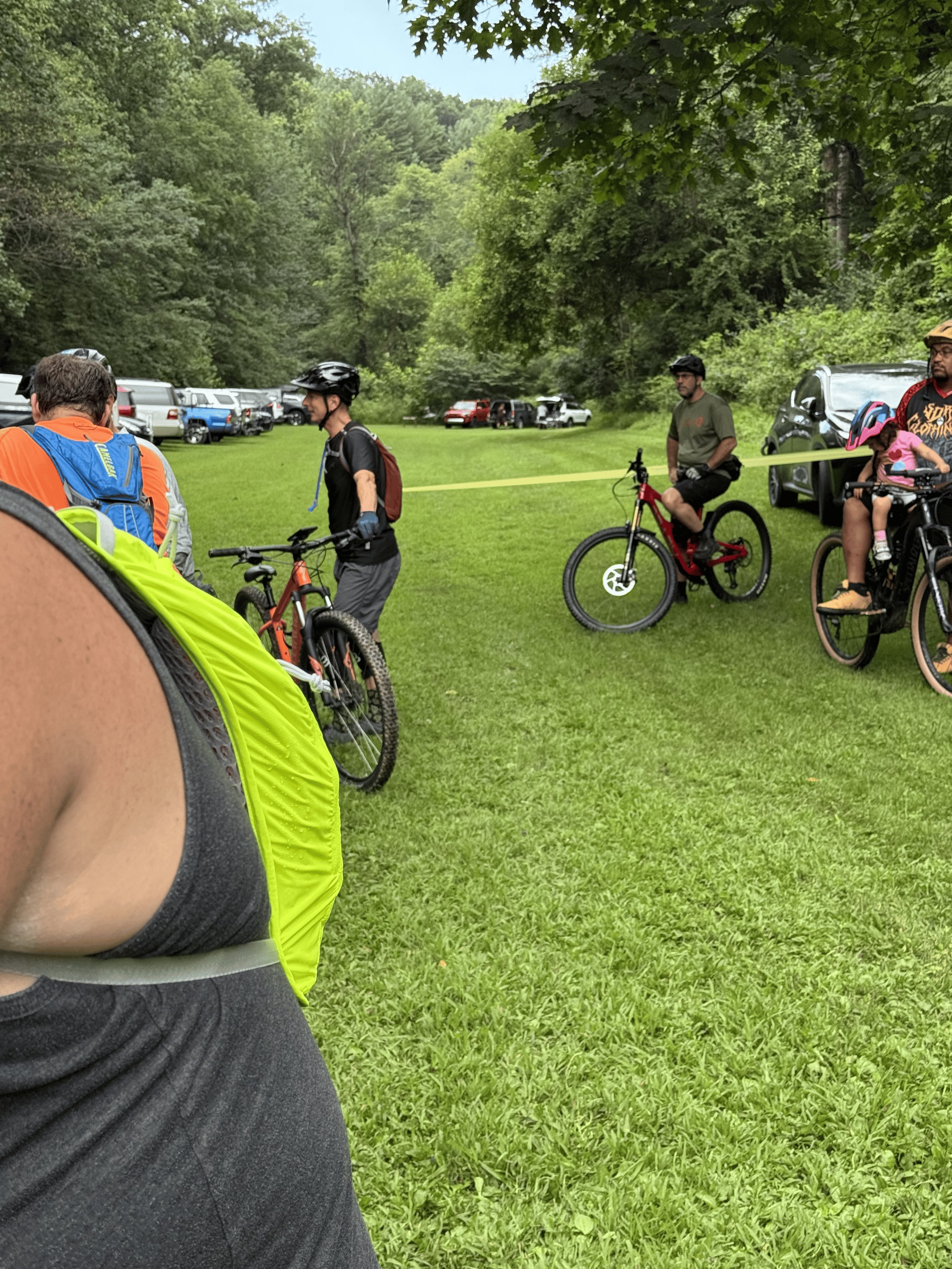 People with bicycles at a park with green grass and trees, cars parked in the background.