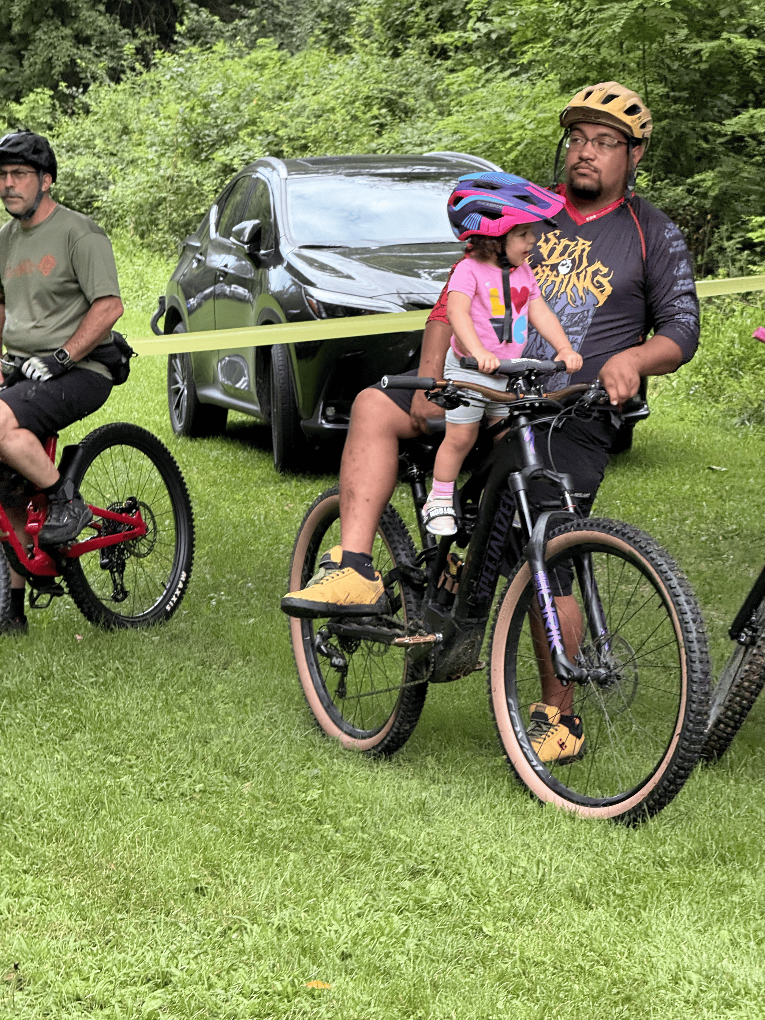 A man and a young girl riding bikes on a grassy area with trees and a black car in the background. The girl is wearing a pink shirt and helmet, while the man is wearing a black long-sleeve shirt, tan helmet, and glasses.