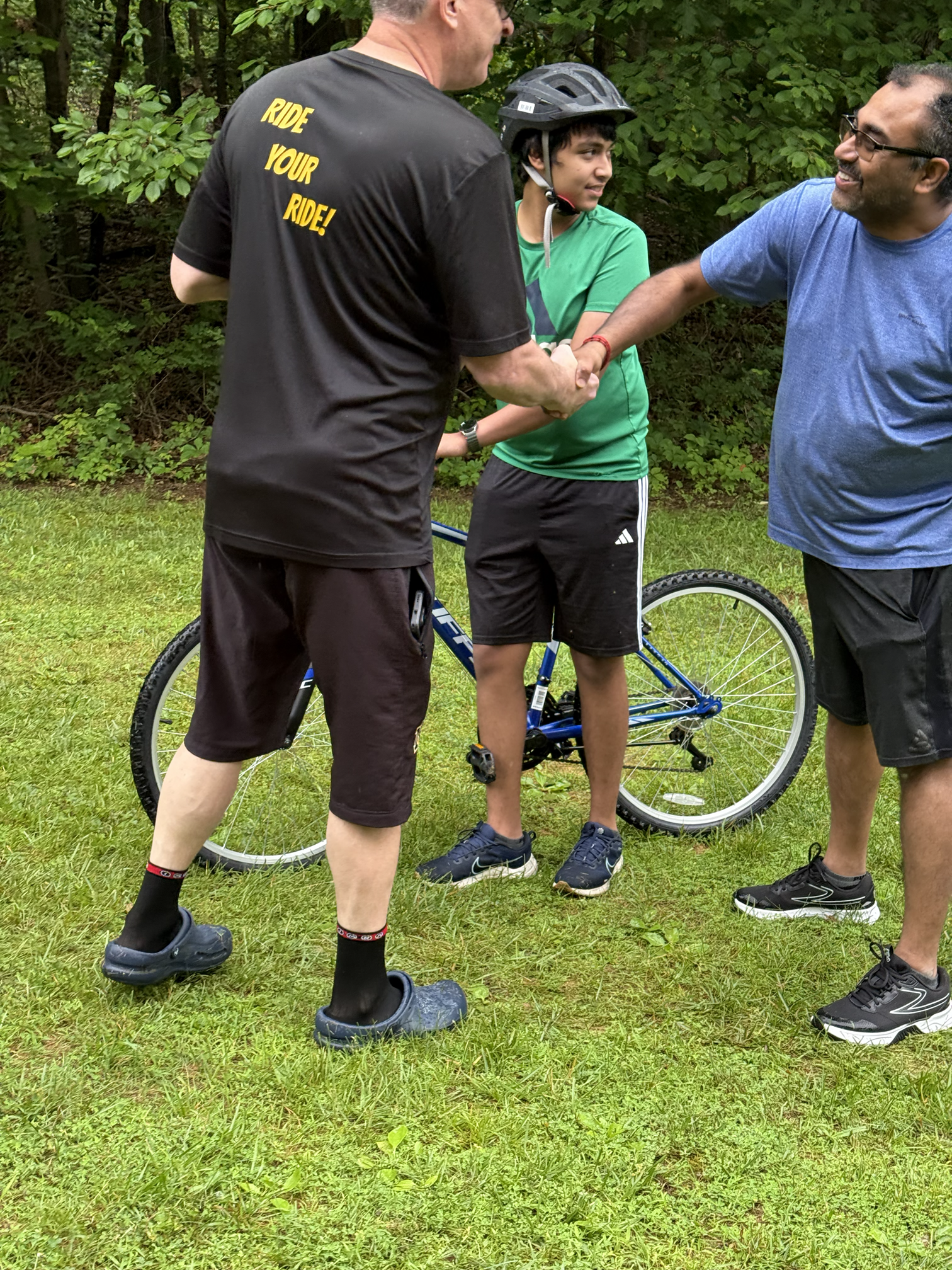Three people outdoors on a grassy area surrounded by trees, engaging in a handshake; one person wears a black t-shirt with yellow text, another is in a green shirt with a bicycle, and the third wears a blue shirt and glasses.