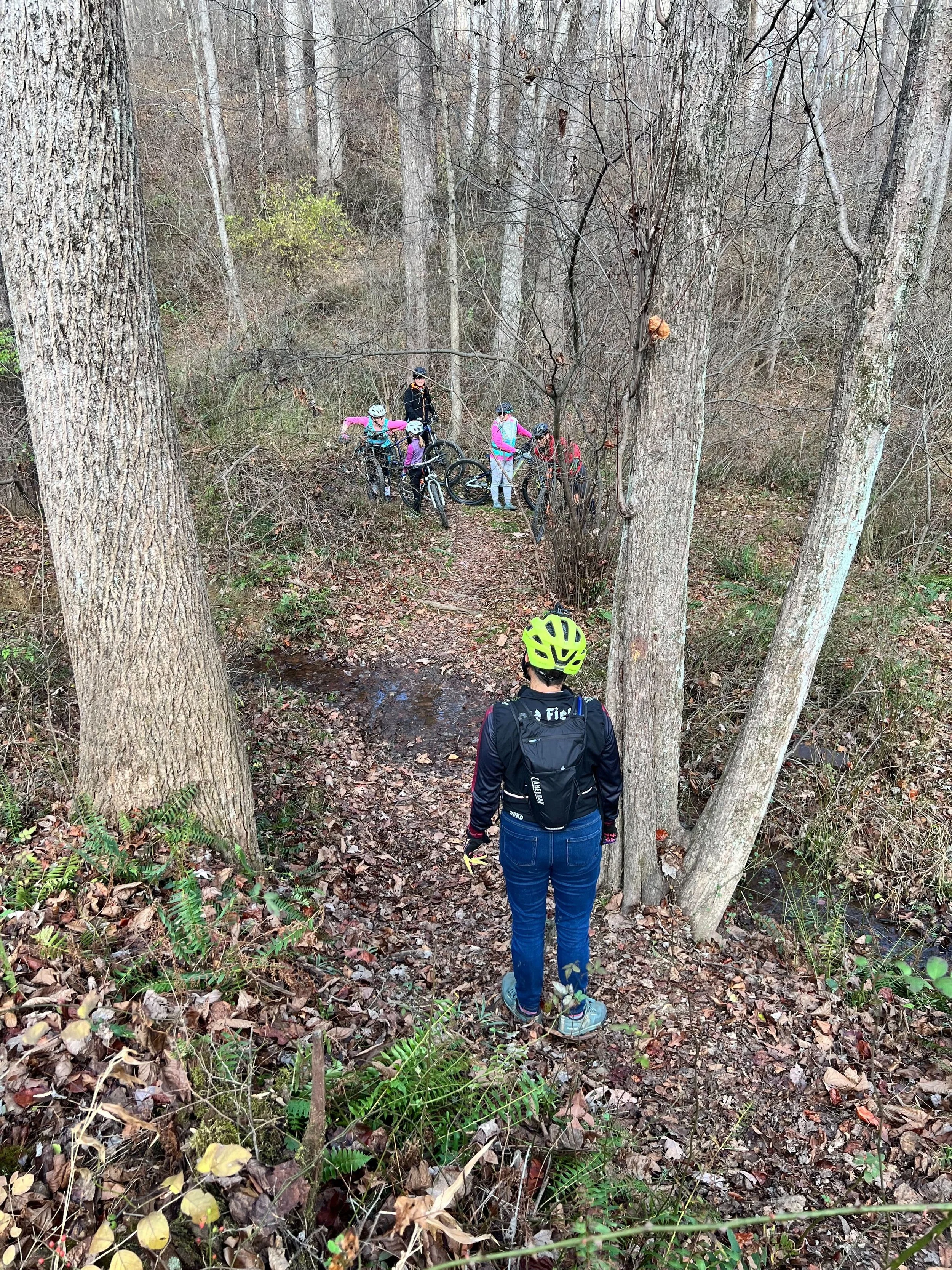 A group of mountain bikers with helmets and gear gather in a wooded area near a small stream, some standing with bikes, others sitting or bending down, surrounded by trees and fallen leaves.