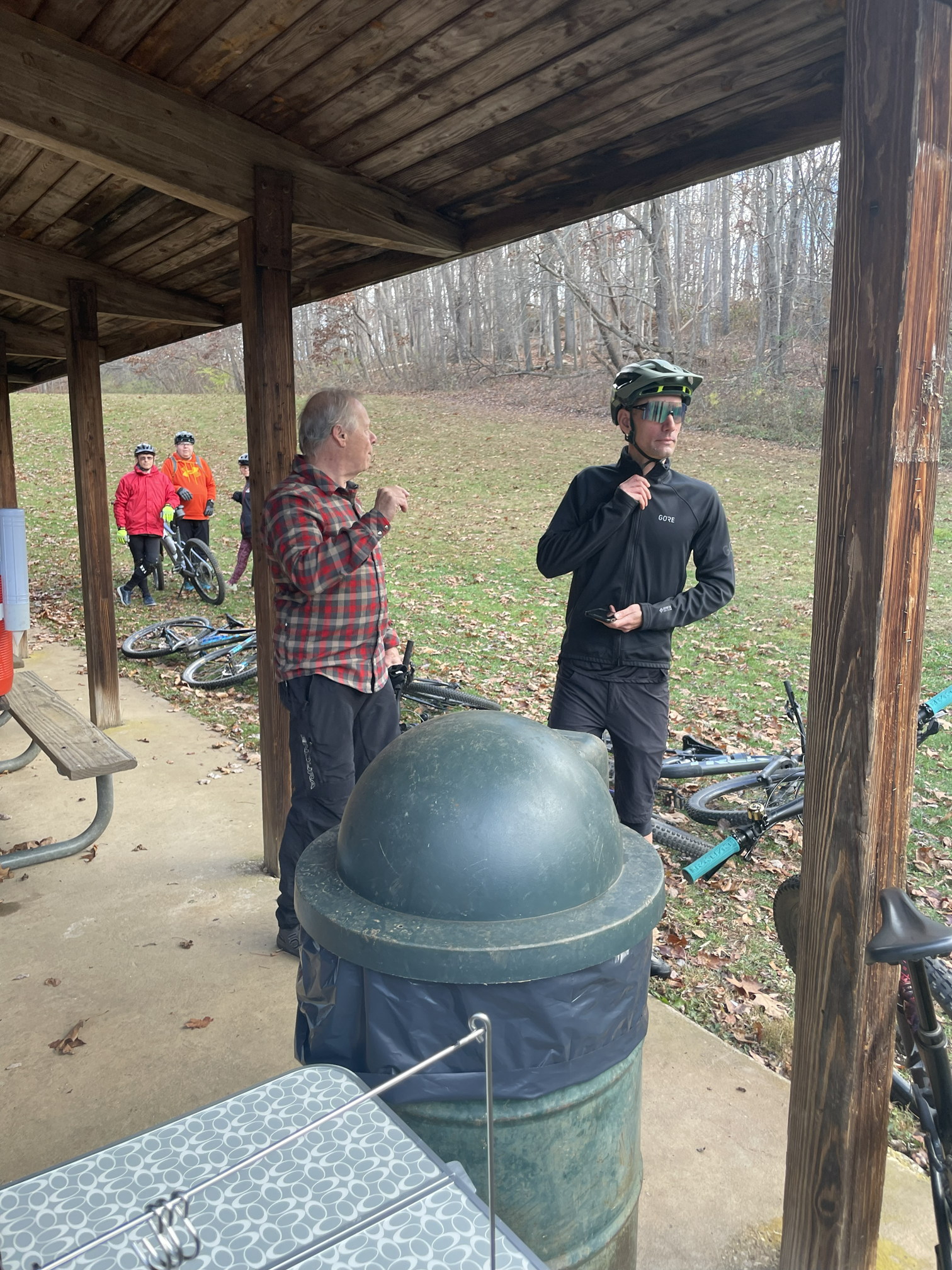 A group of cyclist standing under a wooden shelter, with bicycles lying on the ground nearby. The area appears to be park-like with trees in the background and fallen leaves on the ground.