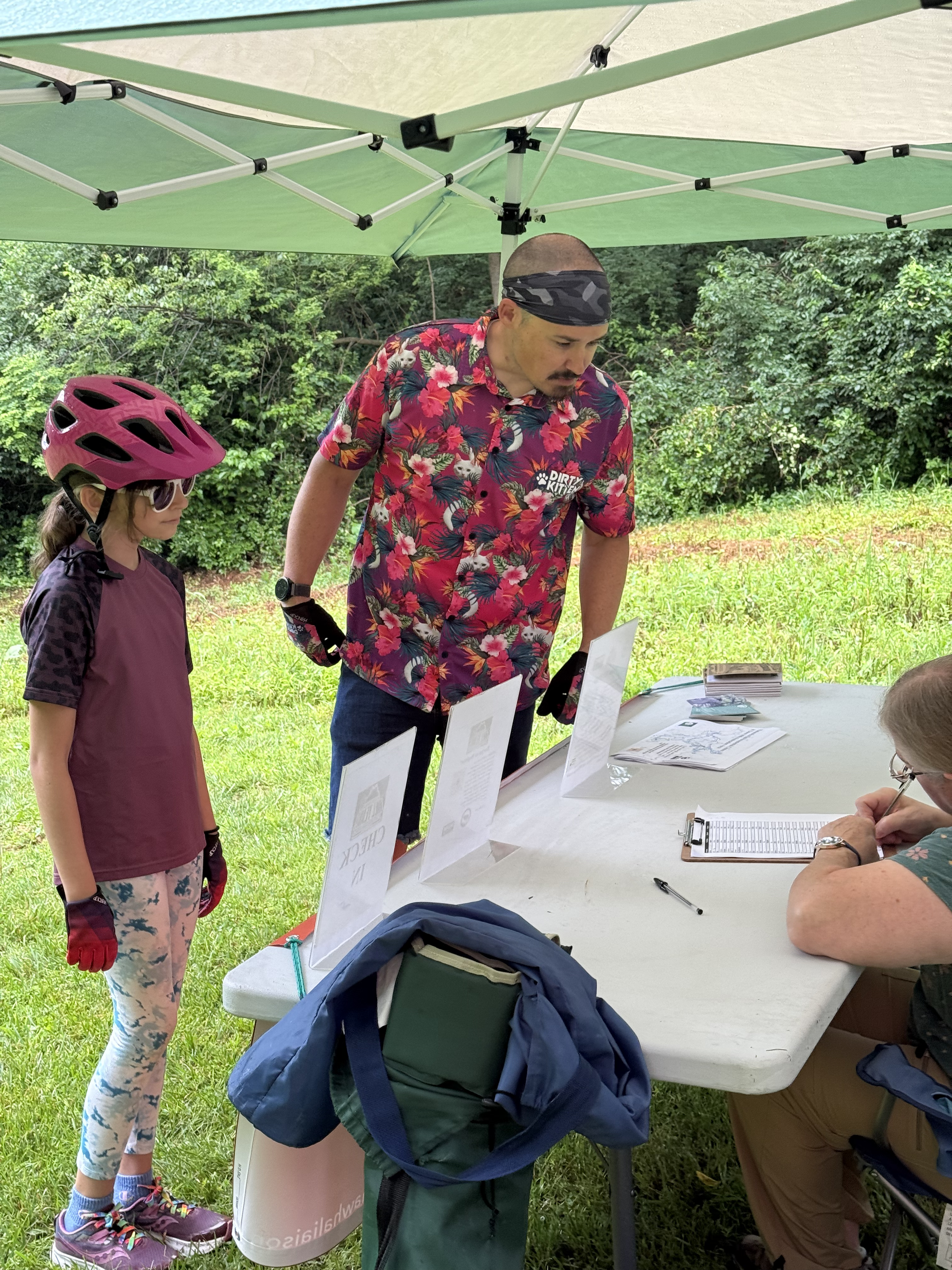 A man and a young girl stand under a green canopy, looking at papers on a table. The man wears a colorful floral shirt, a black wristwatch, and a black headband. The girl wears a purple cycling helmet, sunglasses, a maroon athletic shirt, patterned l