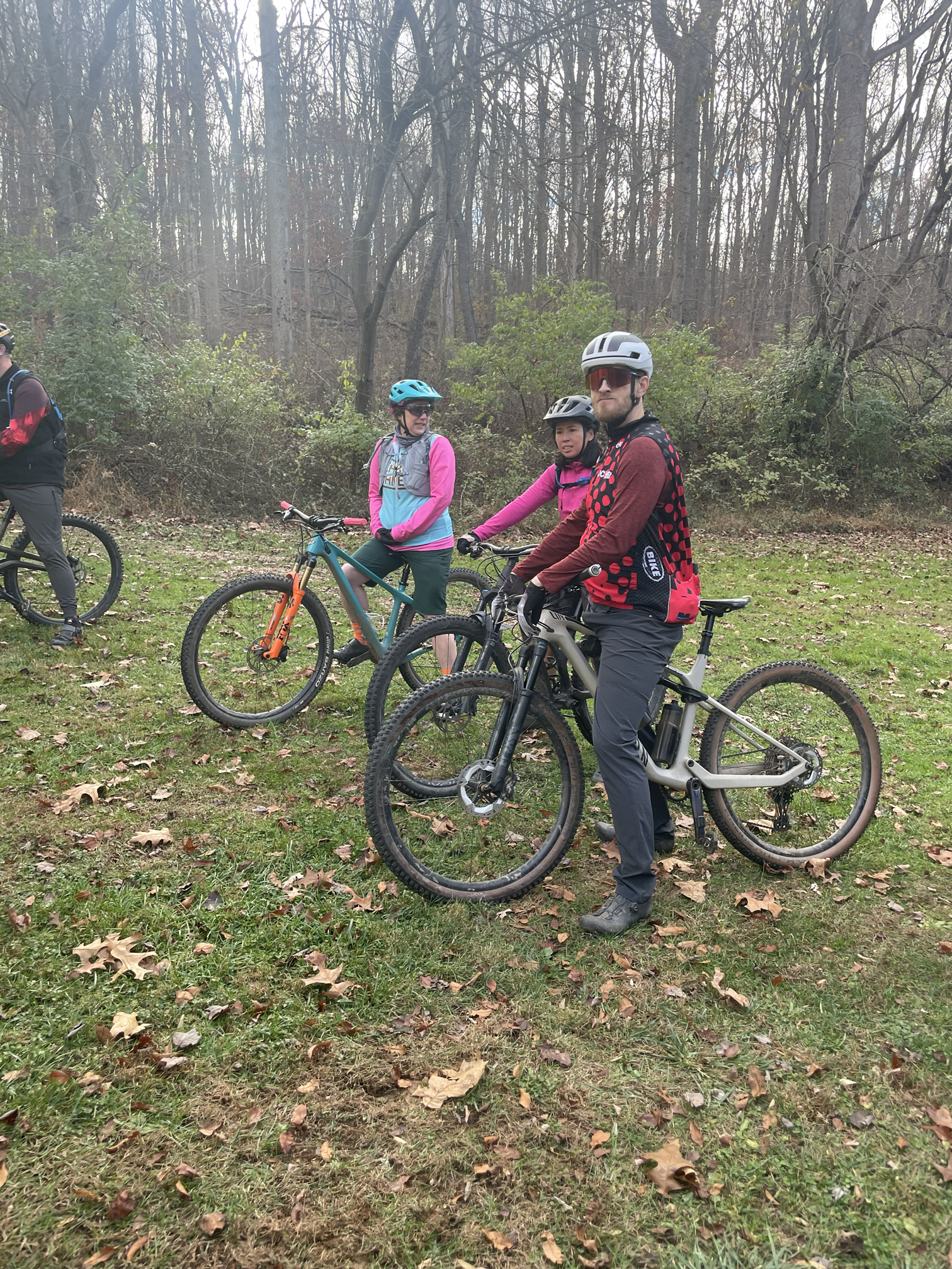 Group of four cyclists with helmets on a grassy trail in a wooded area, wearing outdoor cycling gear.