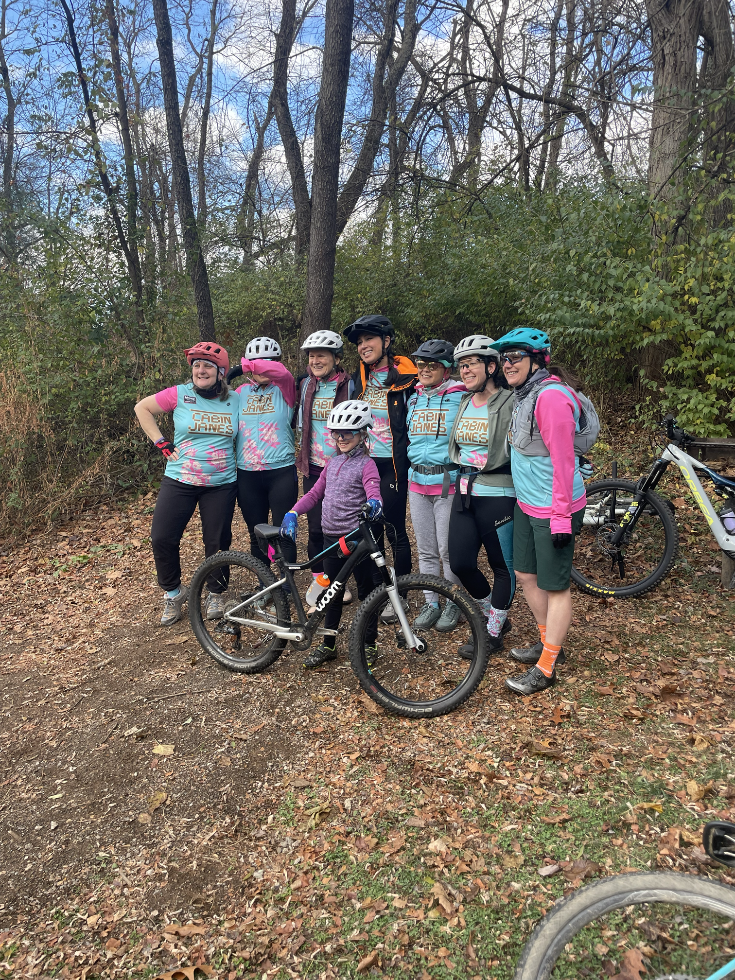 A group of eight women and one young girl posing for a photo on a forest trail, all wearing helmets and cycling gear. They are smiling and standing with their mountain bikes among trees with autumn leaves on the ground.
