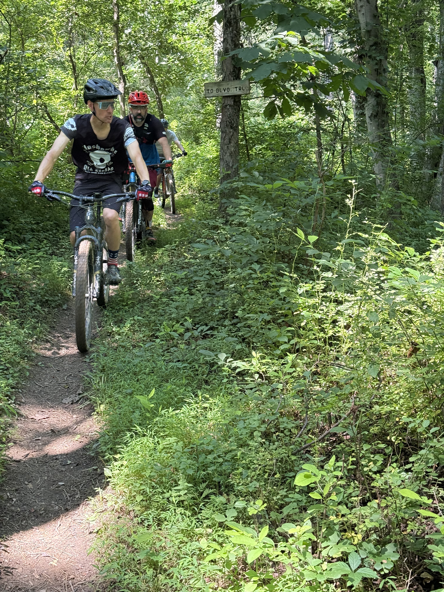 Two men riding mountain bikes on a narrow dirt trail surrounded by dense green foliage and trees in a forest. One is wearing a black helmet and sunglasses, and the other a red helmet.