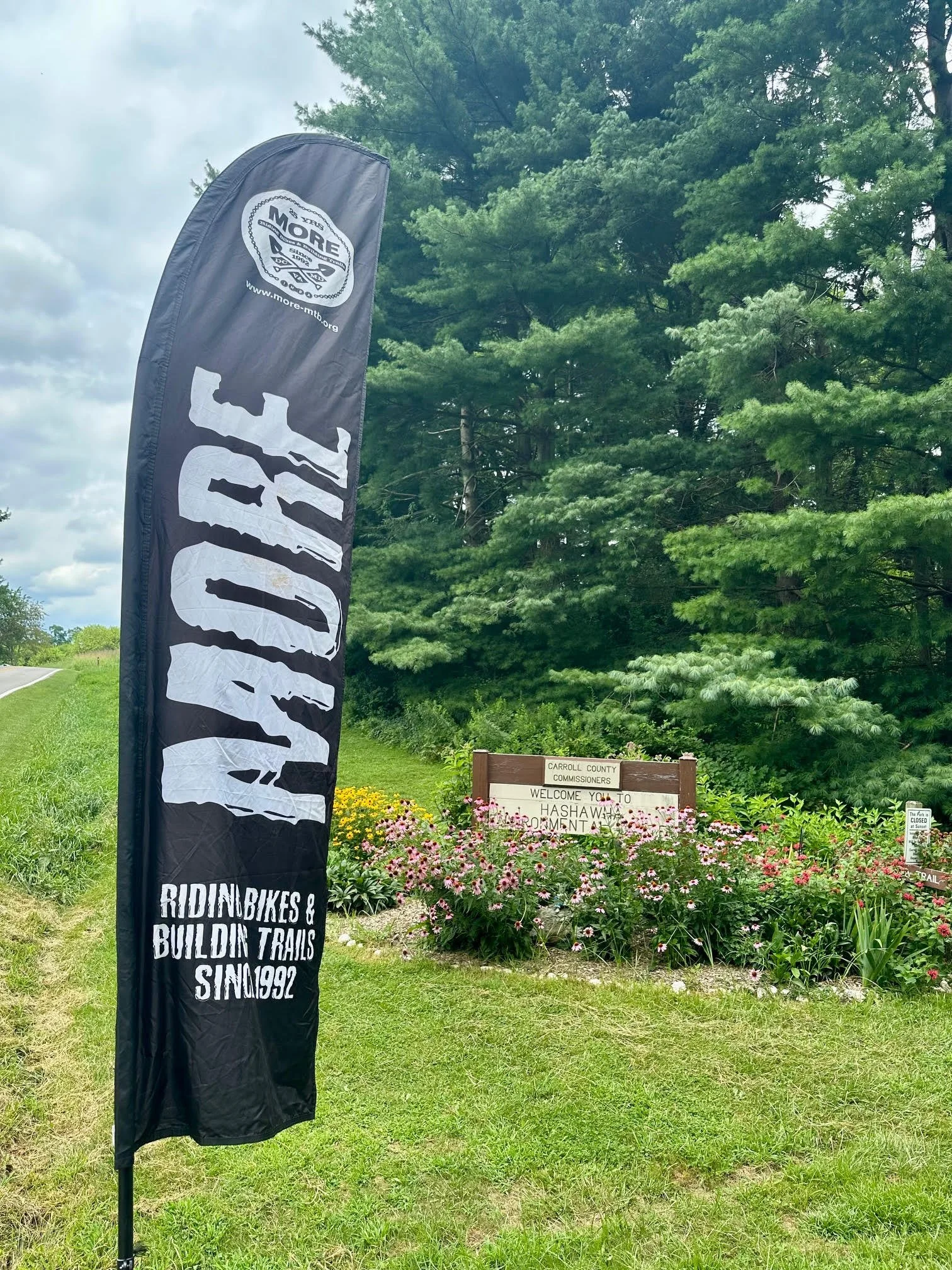 A black vertical banner with white text that says 'RIDE BIKES & BUILDIN TRAILS SINCE 1992', standing on a grassy area near a garden with pink flowers and greenery. In the background, there is a sign that reads 'Carroll County Commissioners Welcome Yo