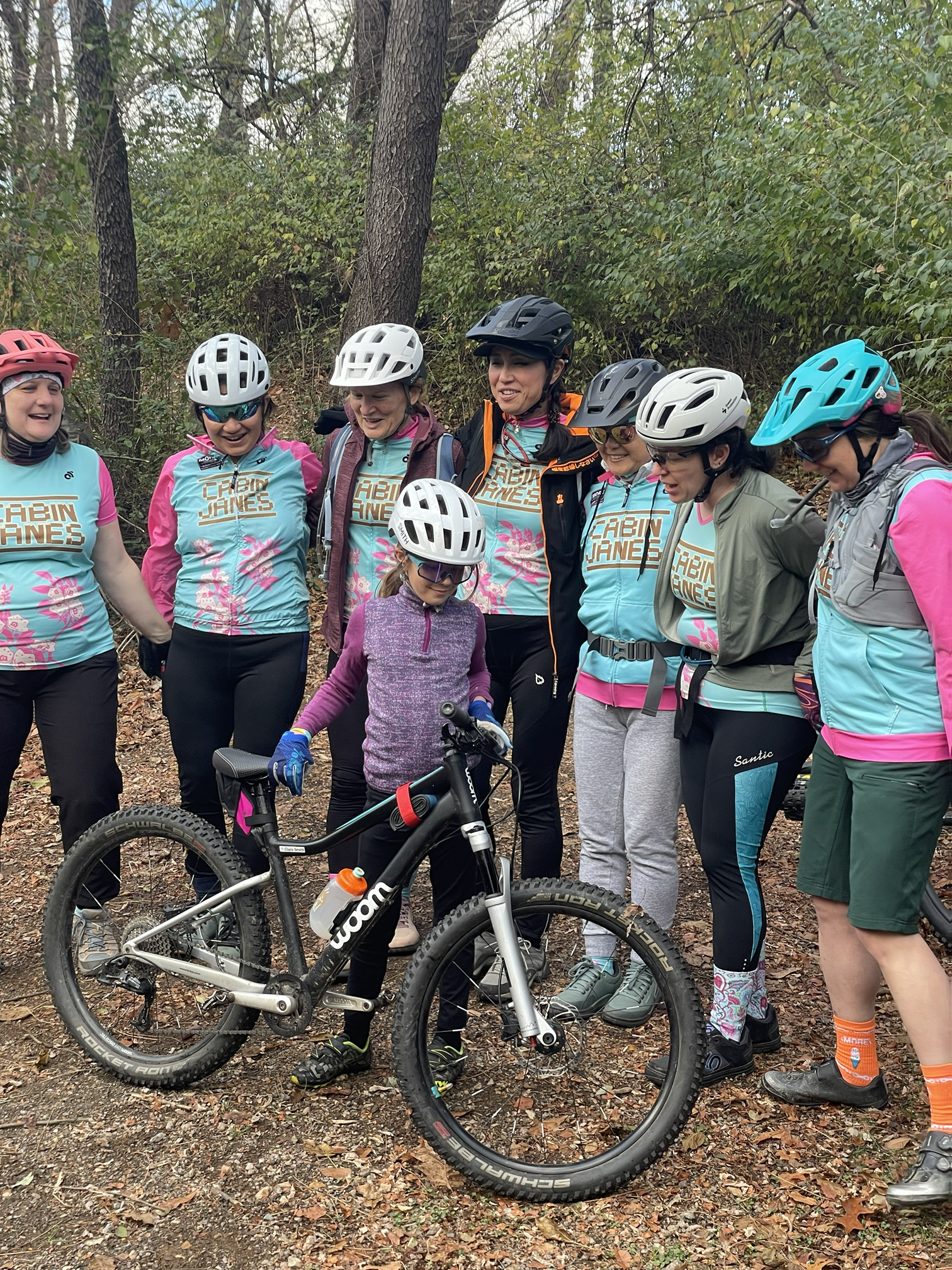 Group of women and a girl in biking gear with helmets, standing outdoors on a forest trail, smiling, with a mountain bike in front of them.