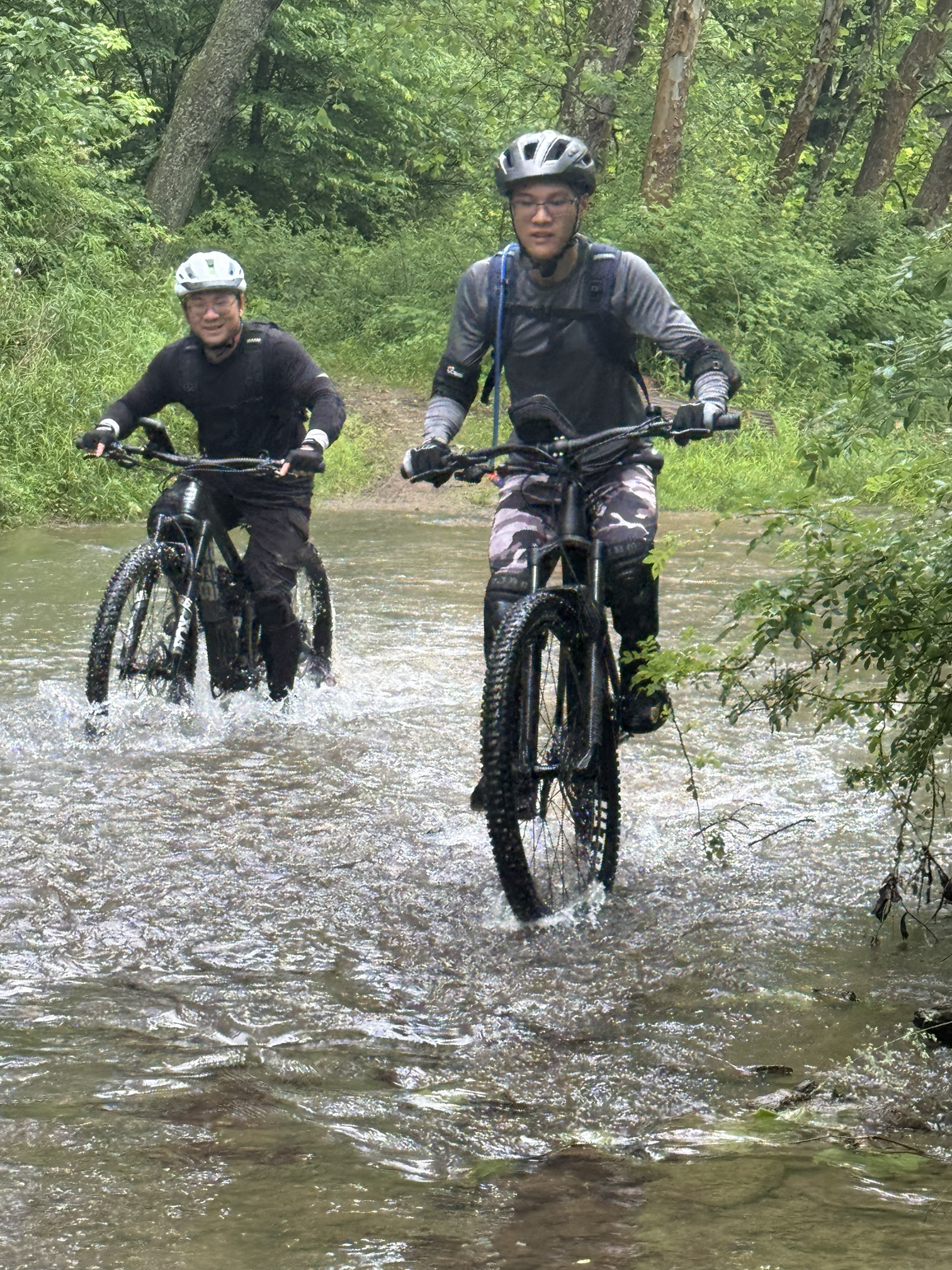 Two mountain bikers wearing helmets and gloves riding through a shallow, rocky stream in a lush green forest.