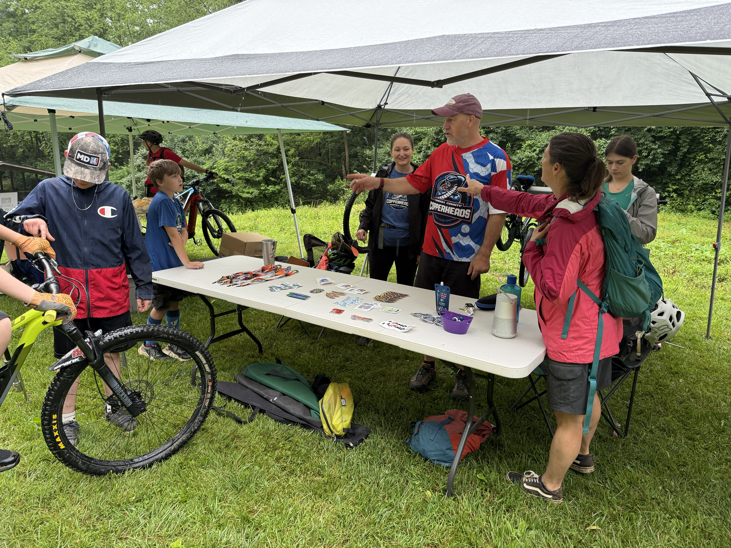 A group of people at an outdoor event under a canopy tent, with bikes and recreational gear visible. The scene occurs in a grassy, wooded area during daytime.